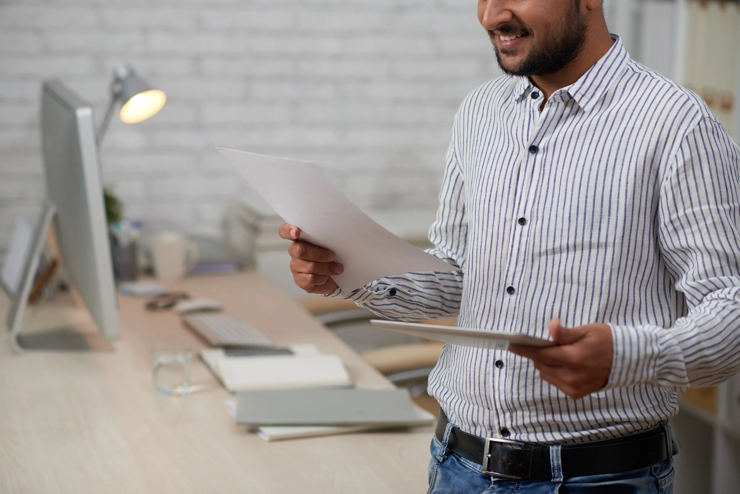 A man in a striped dress shirt holding papers in an office with a computer, desk lamp, and workspace in the background.