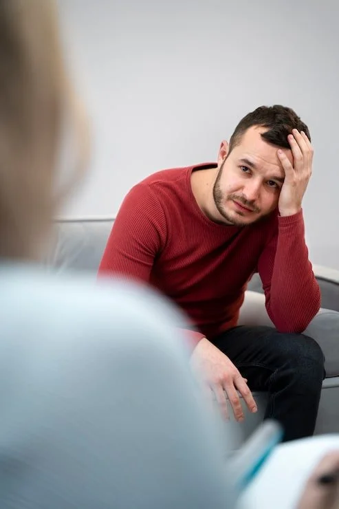 A man sitting on a couch, resting his head on his hand, with a worried or confused expression.