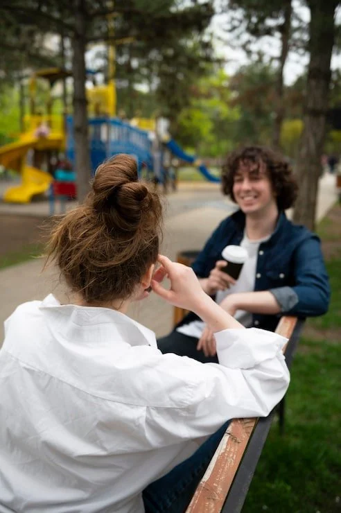 Two people sitting on a park bench, smiling and holding ice cream cones, with a playground in the background.