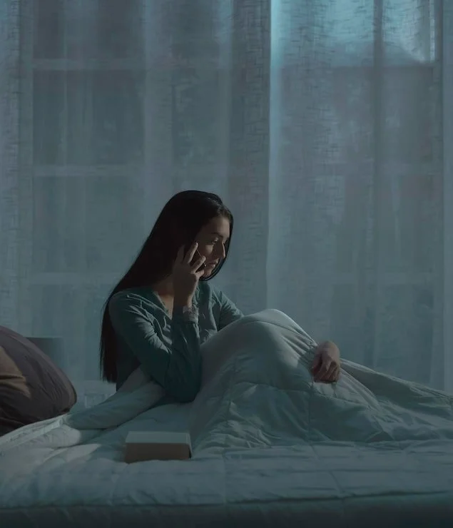 A woman sitting in bed, talking on the phone, with a white book or box on the bed in front of her, in a dimly lit room with sheer curtains and blue light.