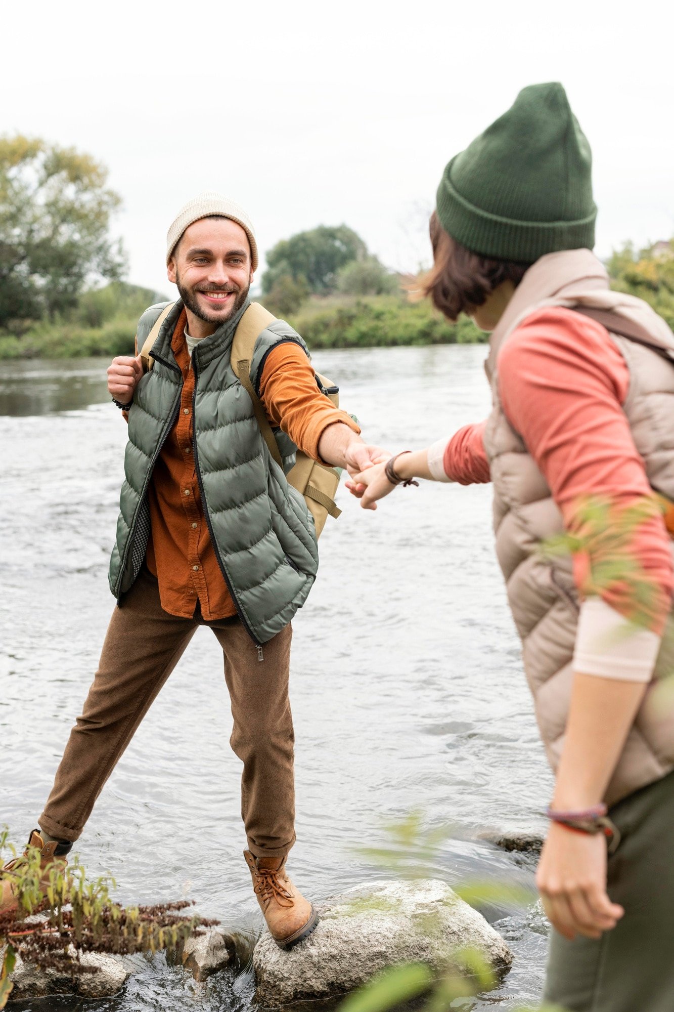 Two people helping each other across a river on rocks. The man is smiling and wearing brown pants, a gray puffer vest, an orange shirt, and a beige beanie. The woman is wearing an orange and tan jacket, green beanie, and glasses.