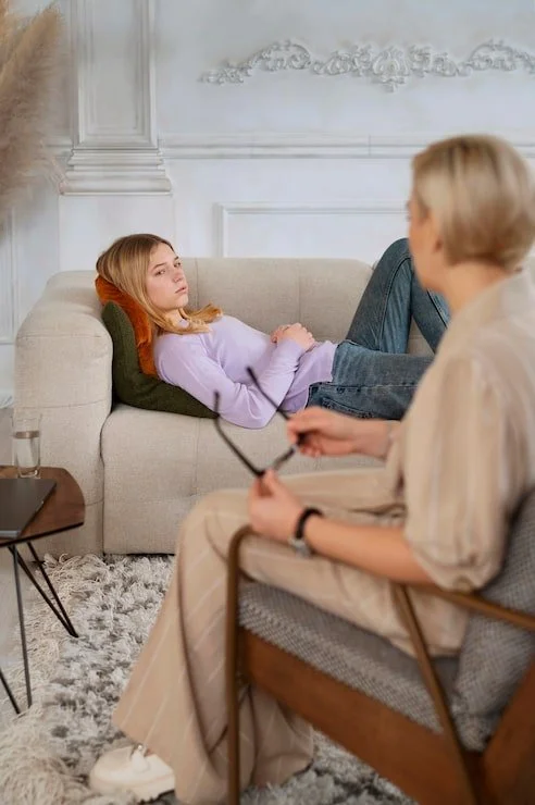 A young girl lies on a beige couch, resting her head on a green pillow, looking thoughtfully at an older woman sitting nearby. The woman is holding glasses and appears to be talking to the girl in a living room with white walls and decorative molding.