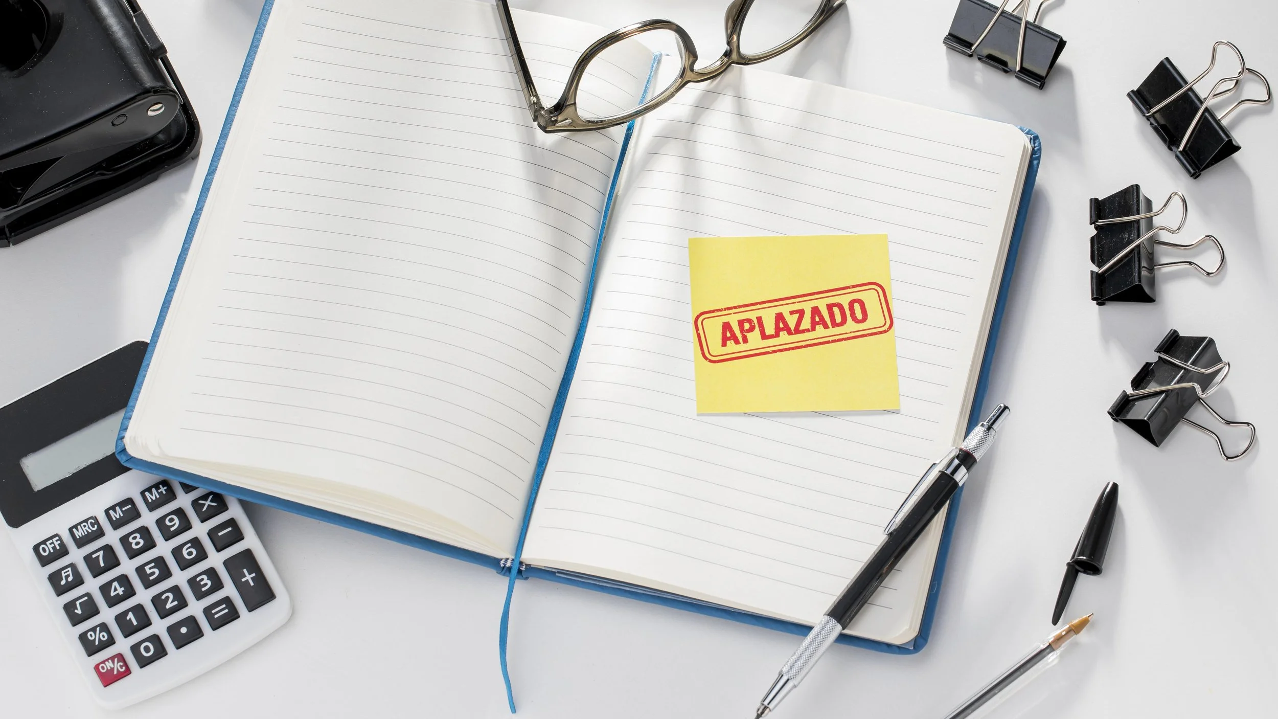 Open notebook with a yellow sticker that says "Aplazado" (Postponed) on a cluttered desk with pens, glasses, a calculator, and binder clips.
