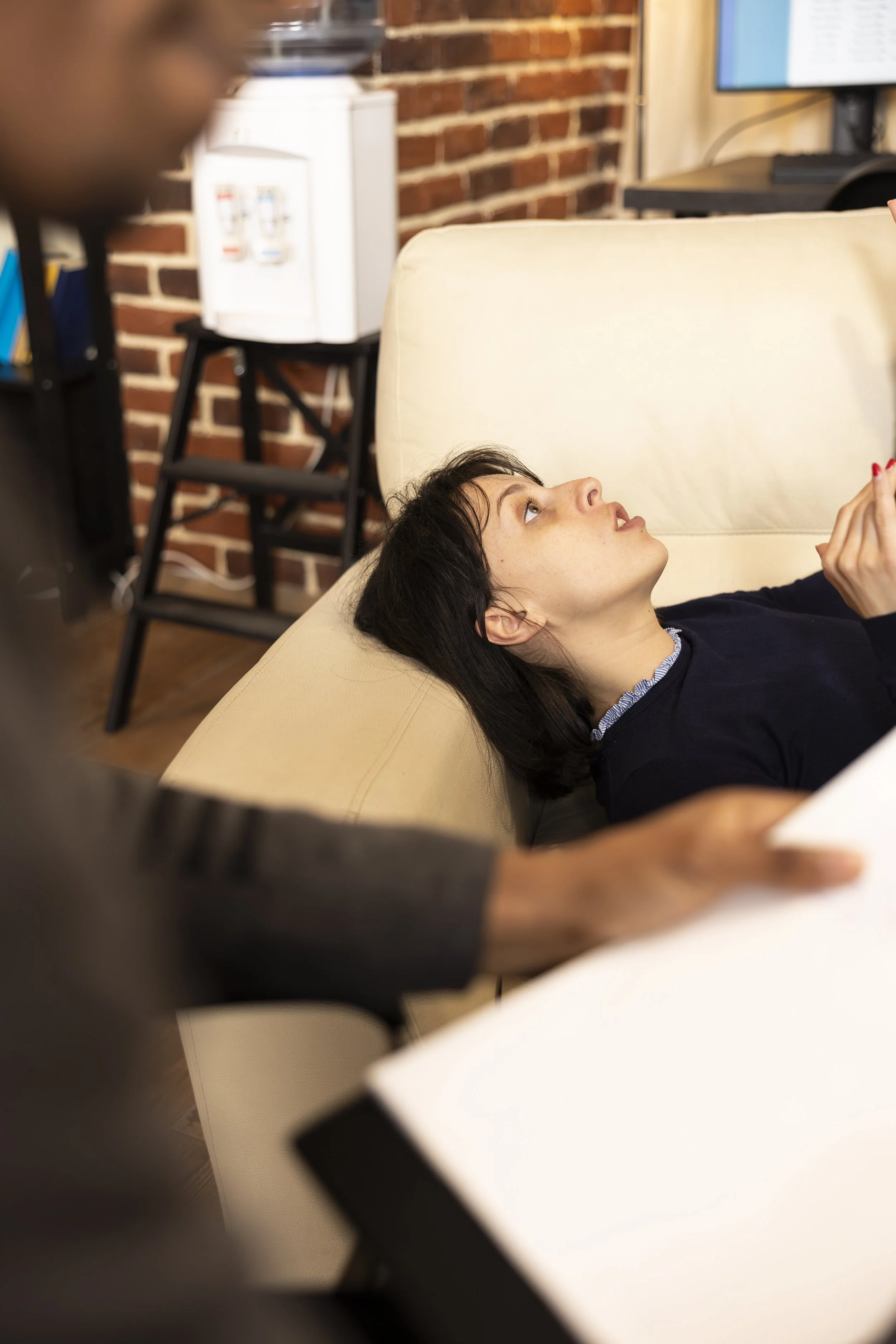 A woman lying on a beige couch looking up, with a man standing in front of her, out of focus. The background features a brick wall, a water dispenser, and a computer monitor.