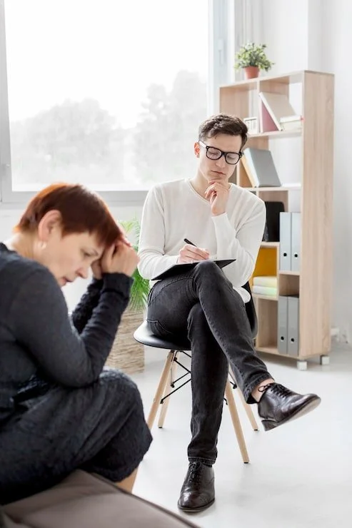 A man and a woman during a counseling session; the woman appears distressed, and the man is taking notes, sitting in a bright room with a bookshelf and a large window.