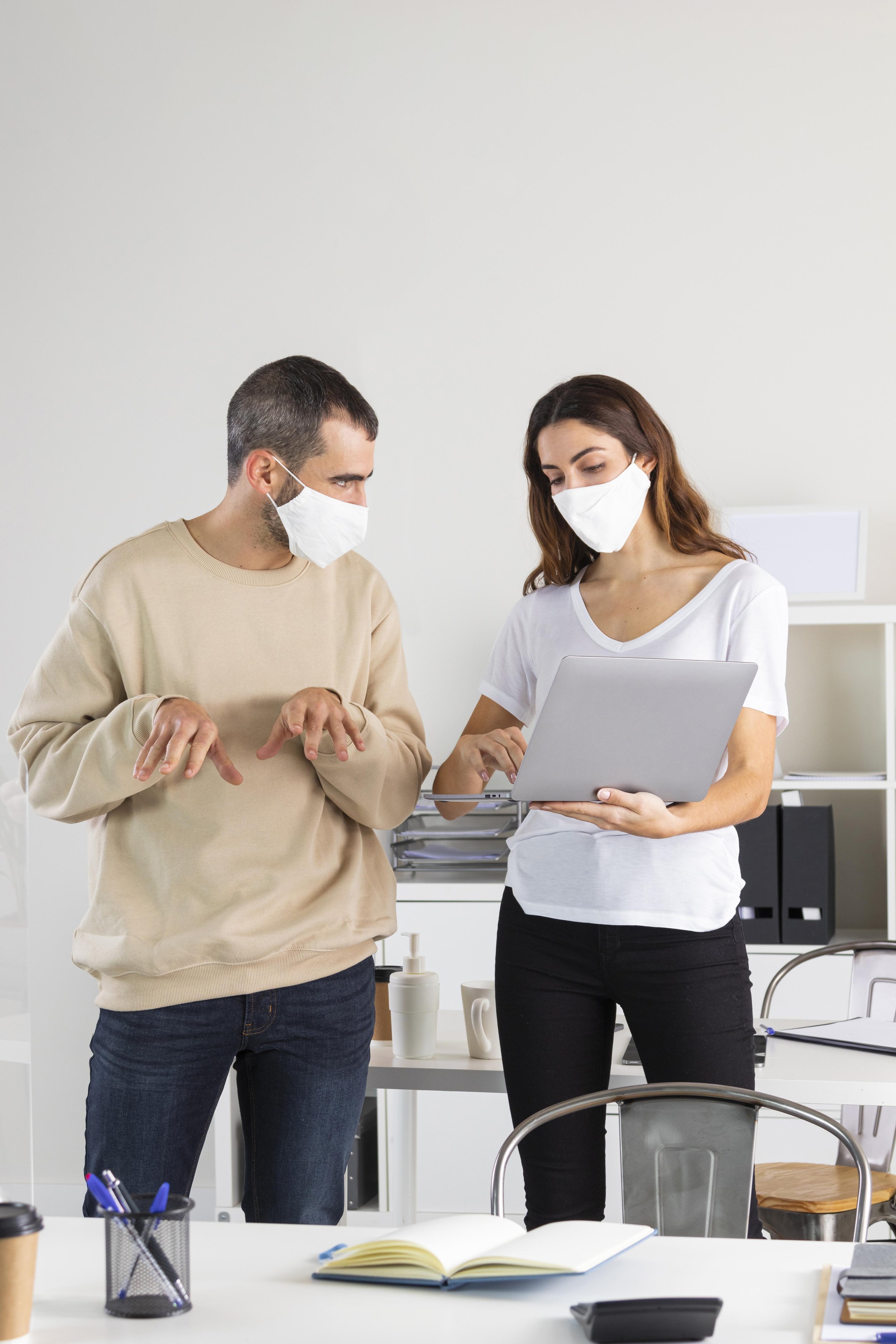 Two people wearing masks, standing in an office, looking at a laptop screen. One is a man with short dark hair and a beige sweater, the other a woman with long brown hair and a white T-shirt. There are office supplies on the desk, including a notebook, pens, and a cup.