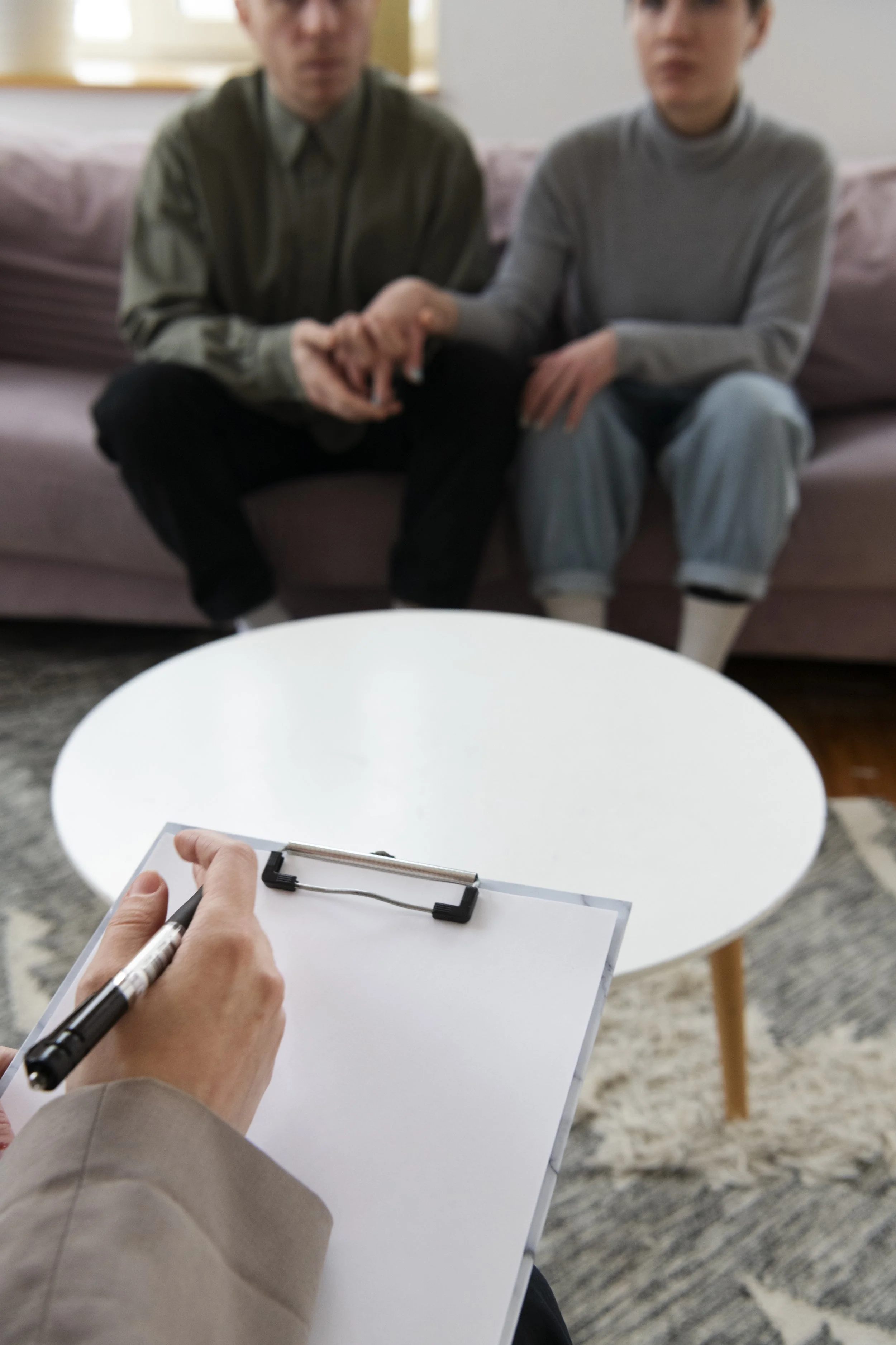 Two individuals sitting on a pink sofa hold hands, while a person in the foreground writes on a clipboard with a clipboard paper and pen. The background includes a light and airy room with large windows.