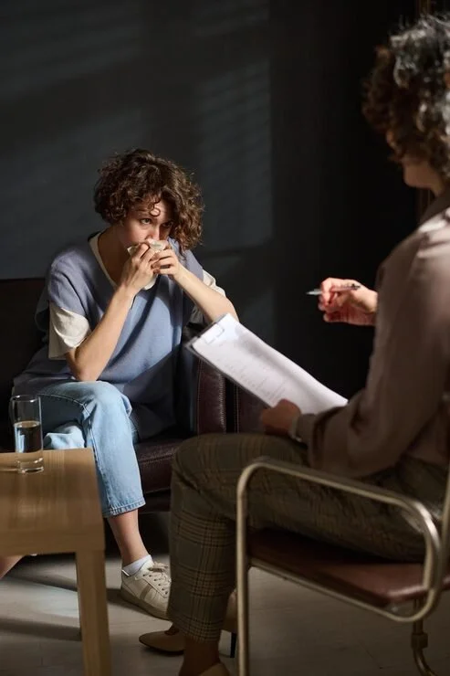 A woman sits on a couch, nervously holding her hands near her face, during a therapy session with a therapist who is taking notes.