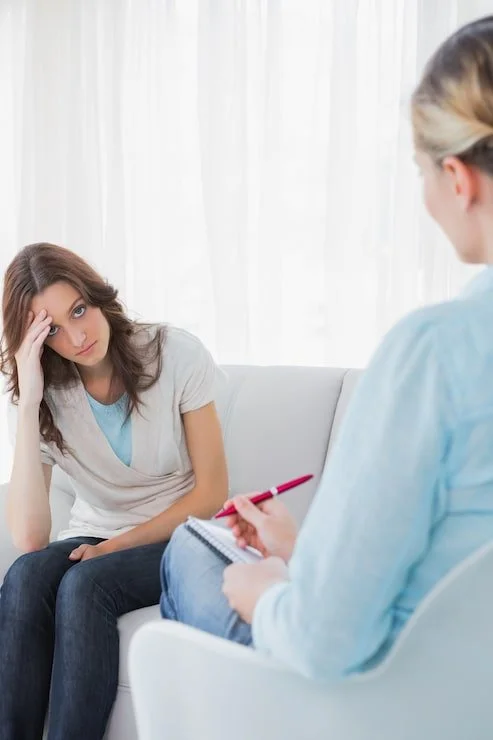 Young woman during therapy session, appearing distressed, sitting on a couch with a therapist taking notes.