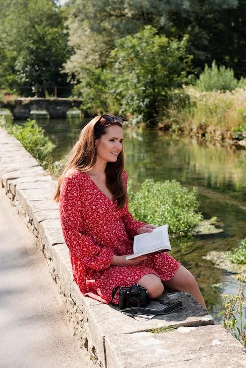 A woman in a red floral dress sits on a stone ledge by a river, holding an open book, with a camera placed beside her, surrounded by green trees and foliage.