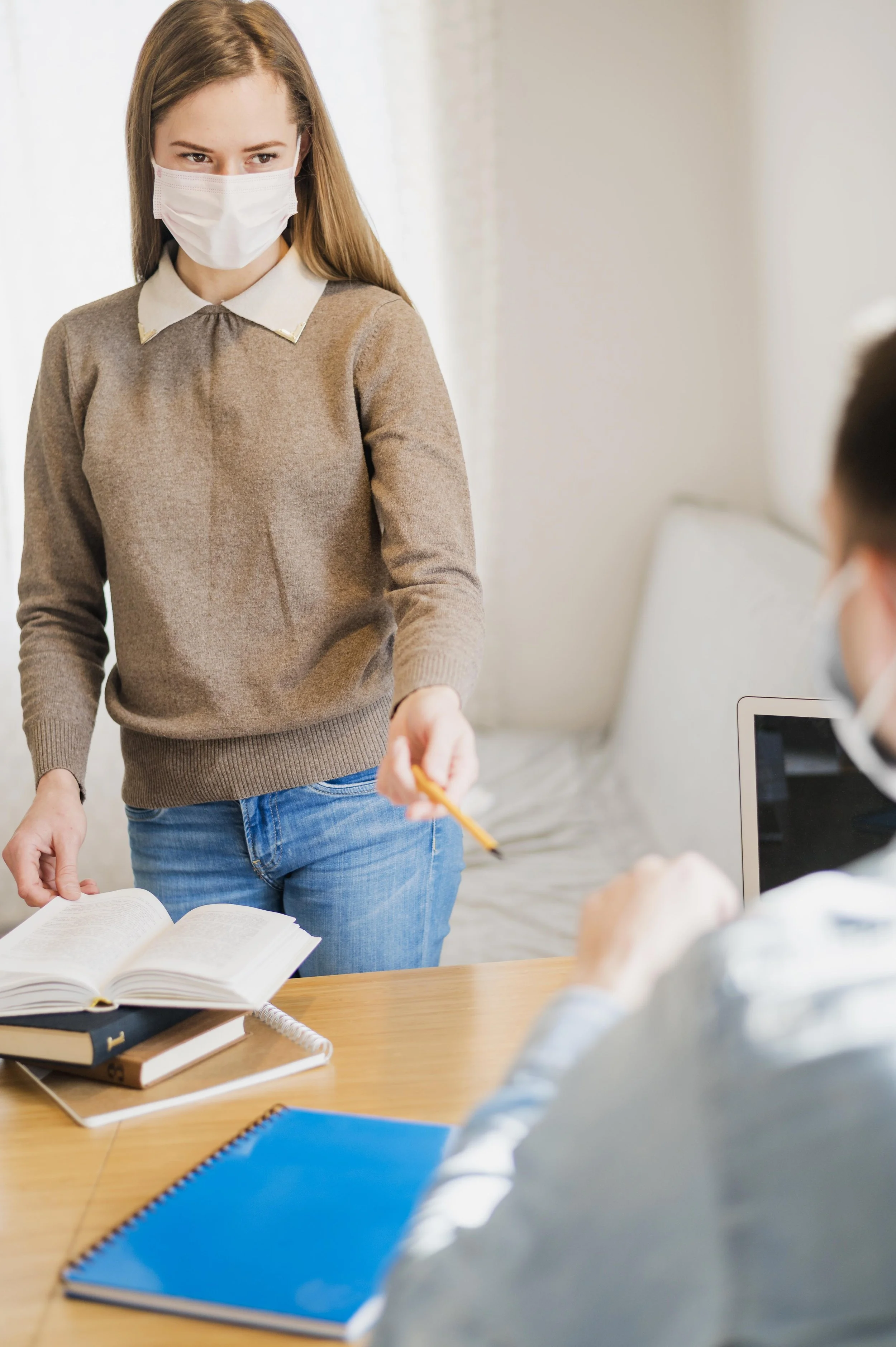 A young woman wearing a face mask, tan sweater with a white collar, and jeans, is handing a yellow pen to a person sitting at a table with books, papers, and a laptop in a classroom or office setting.