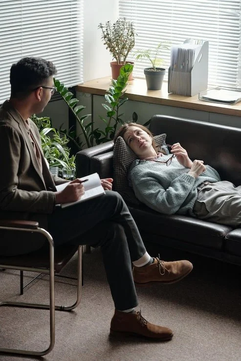 A woman lying on a black couch with glasses in her hand, talking to a man sitting across from her with a notebook. They are in an office with plants and window blinds.