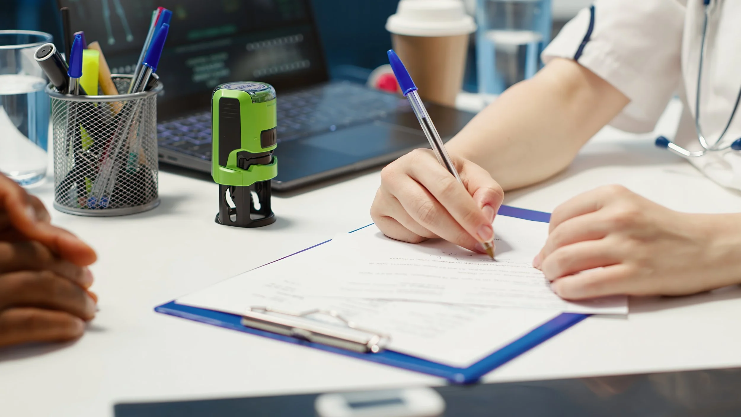 A person in a medical uniform signing a document at a desk in a medical office, with pens, a green device, and a laptop on the desk.