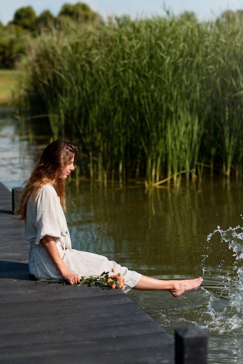A woman sitting on a wooden dock by the water, kicking her foot into the water. She has a bouquet of flowers beside her and tall green reeds in the background.