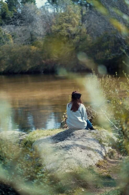 A person with long hair, wearing a light-colored sweater, sitting on a large rock by a river, surrounded by trees and nature.