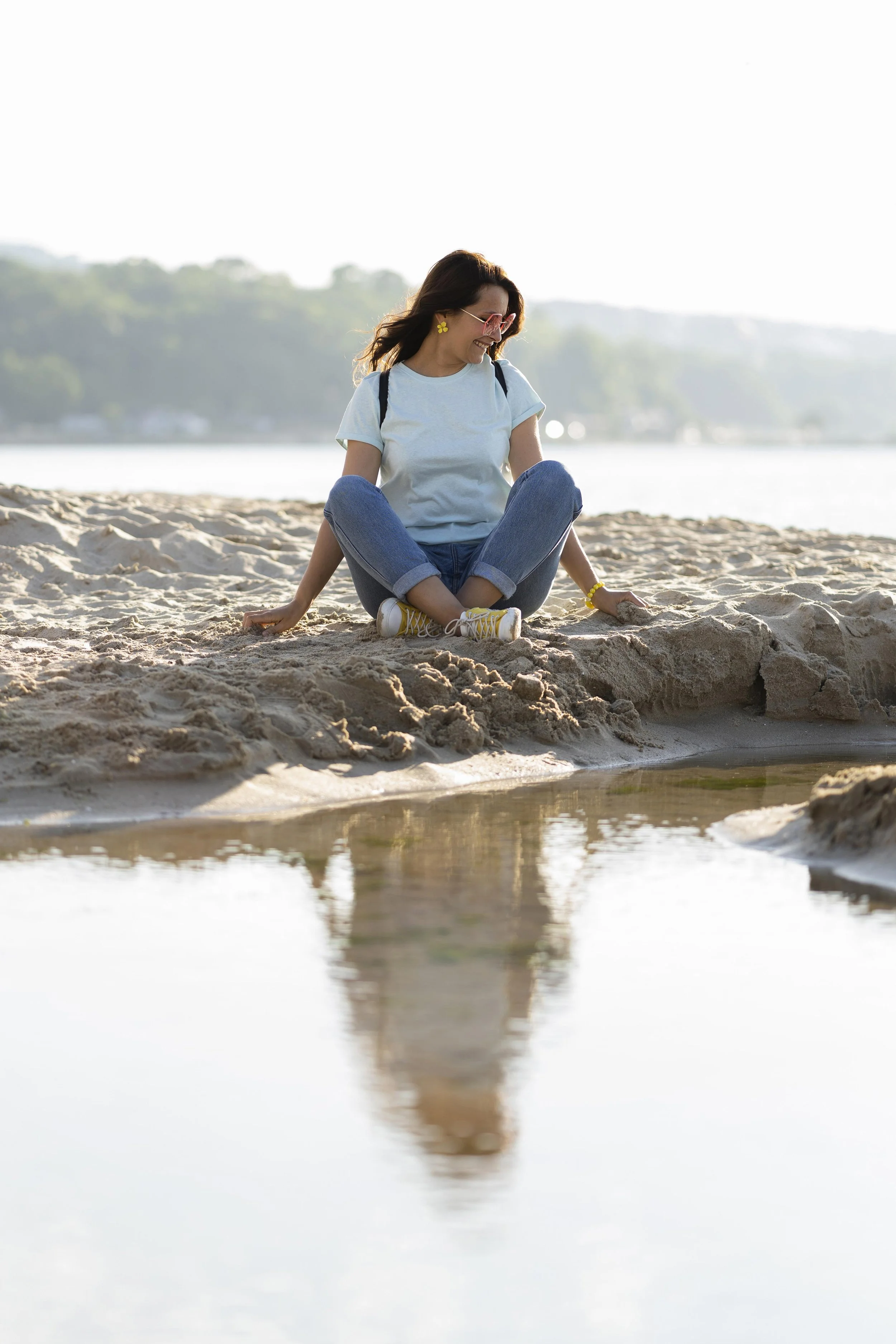 Woman sitting on a sandy beach near a small tide pool, wearing glasses, a light blue t-shirt, jeans, and sneakers. She is smiling and looking down, with a backdrop of water and distant trees.