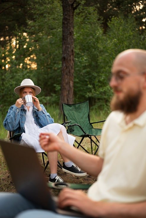 A woman sitting outdoors in a forested area, drinking from a metal mug, wearing a white hat, denim jacket, and white dress. A man in the foreground, slightly blurred, working on a laptop, is holding a small object in his hand. There are camping chairs and trees in the background.