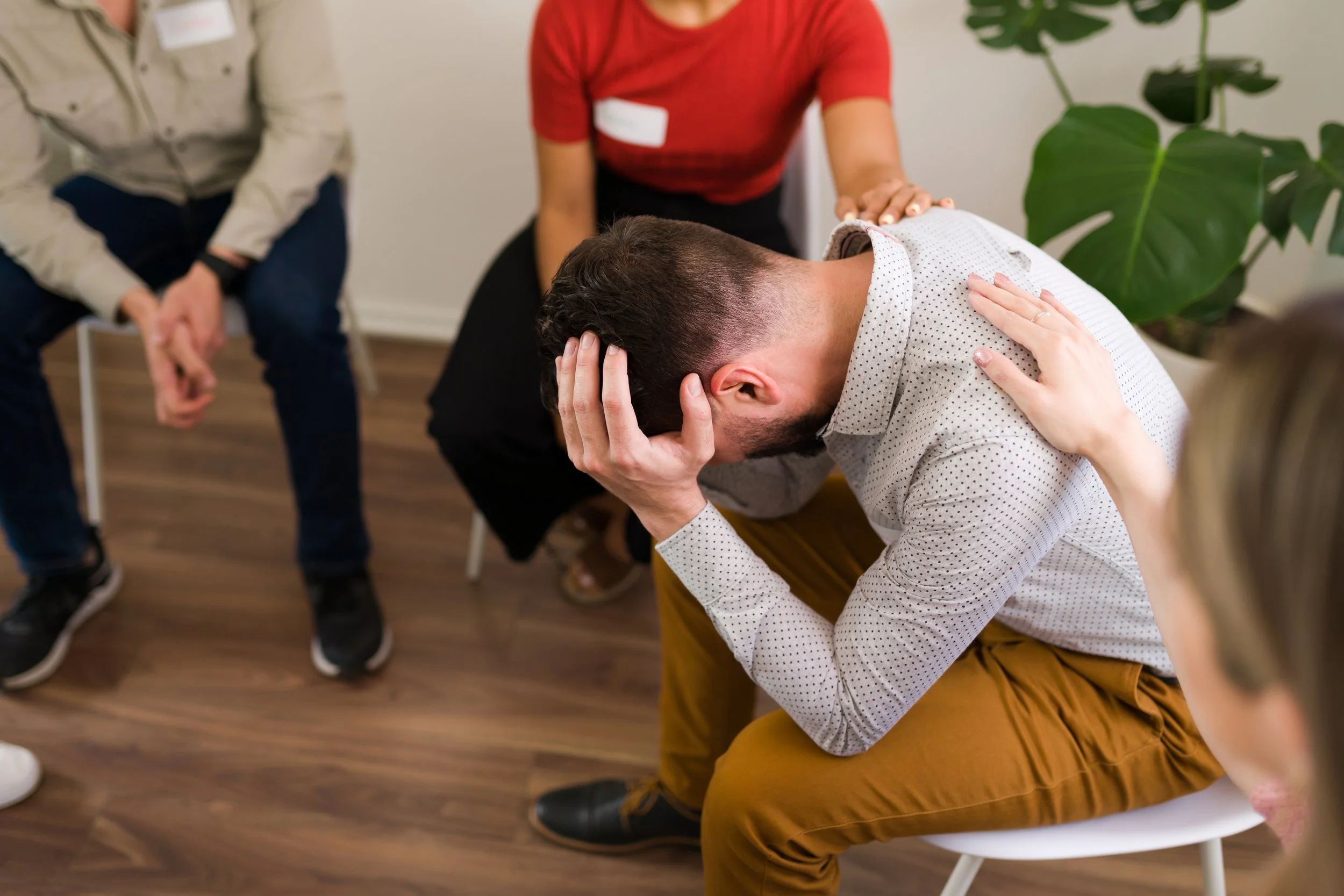 A man sitting with his head bowed and hands clasped on his head, appearing distressed. A woman with a hand on his shoulder is supporting him. Two other people are seated nearby, observing the scene. The setting appears to be a therapy or counseling session in a room with wooden flooring and a large green plant.