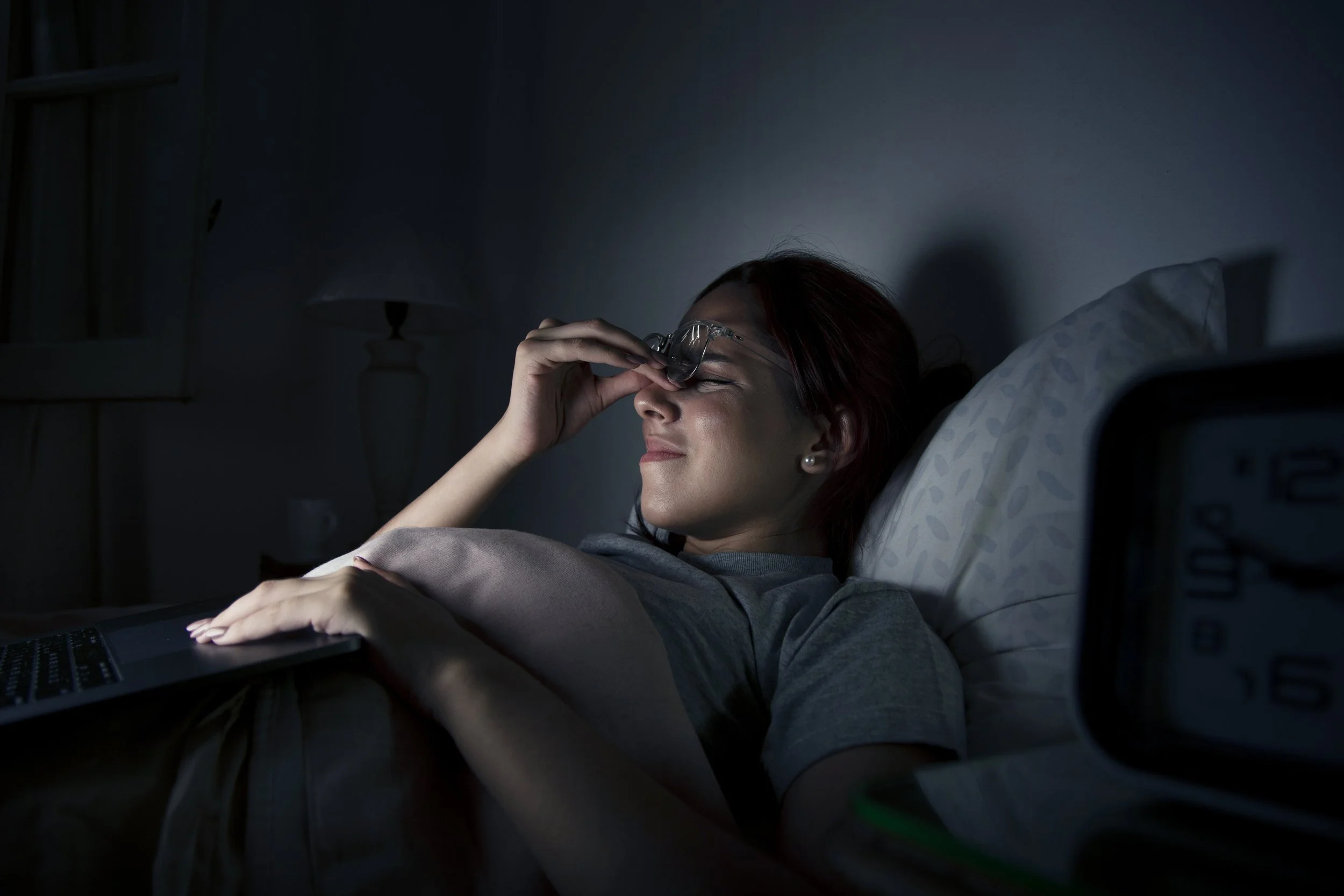 A woman lying in bed with her eyes closed, holding her glasses to her forehead, appearing to be tired or resting, in a dimly lit room.