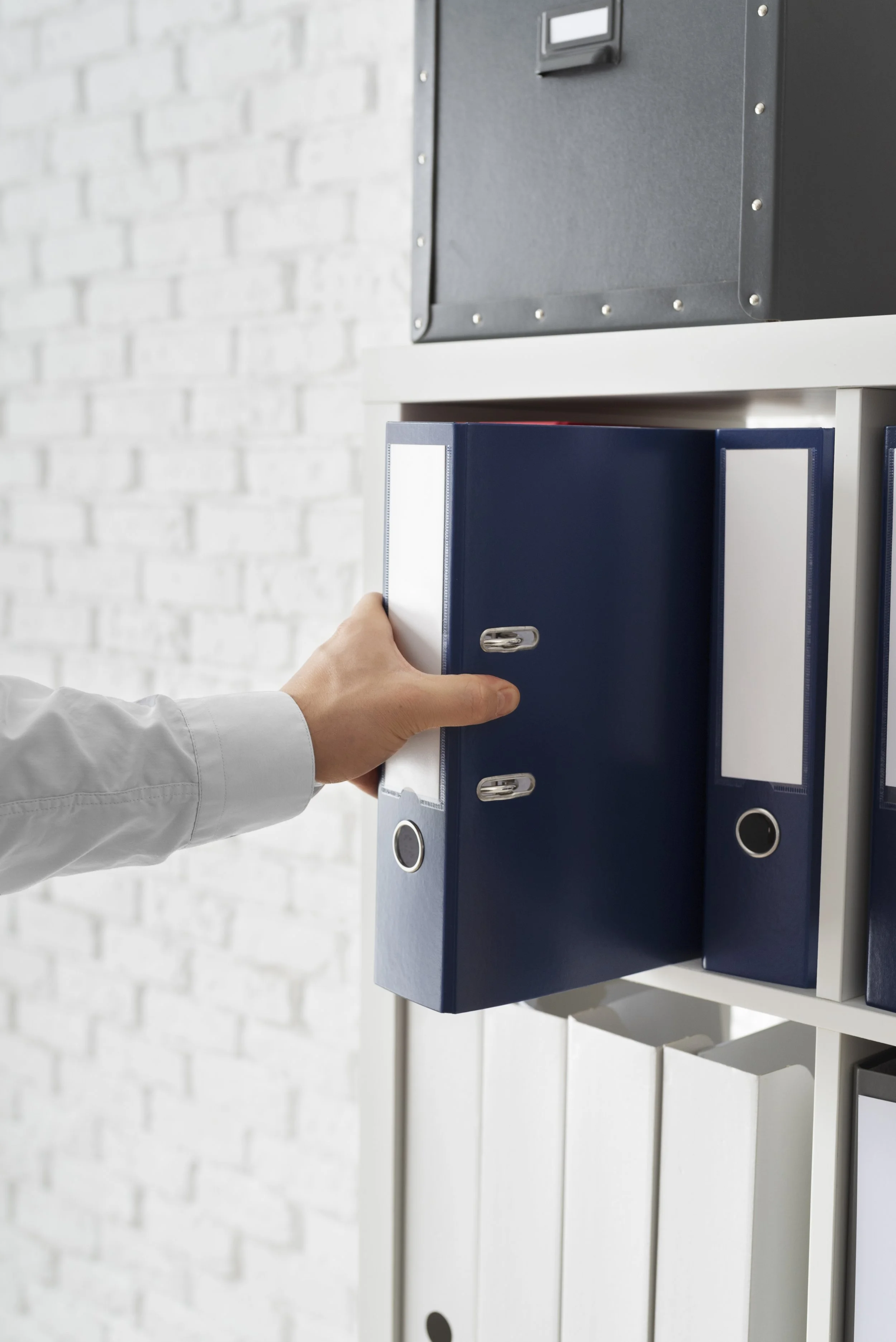 A person's hand placing a blue file folder into a white vertical filing cabinet, with a mounted black electronic device or safe on the wall behind.