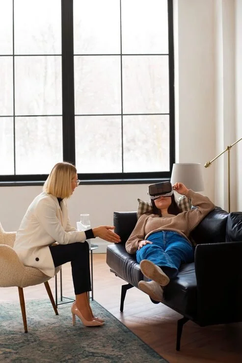 A woman is sitting on a couch wearing a virtual reality headset, with a woman sitting on a chair in front of her, talking and holding a glass of water, in a bright room with large windows.