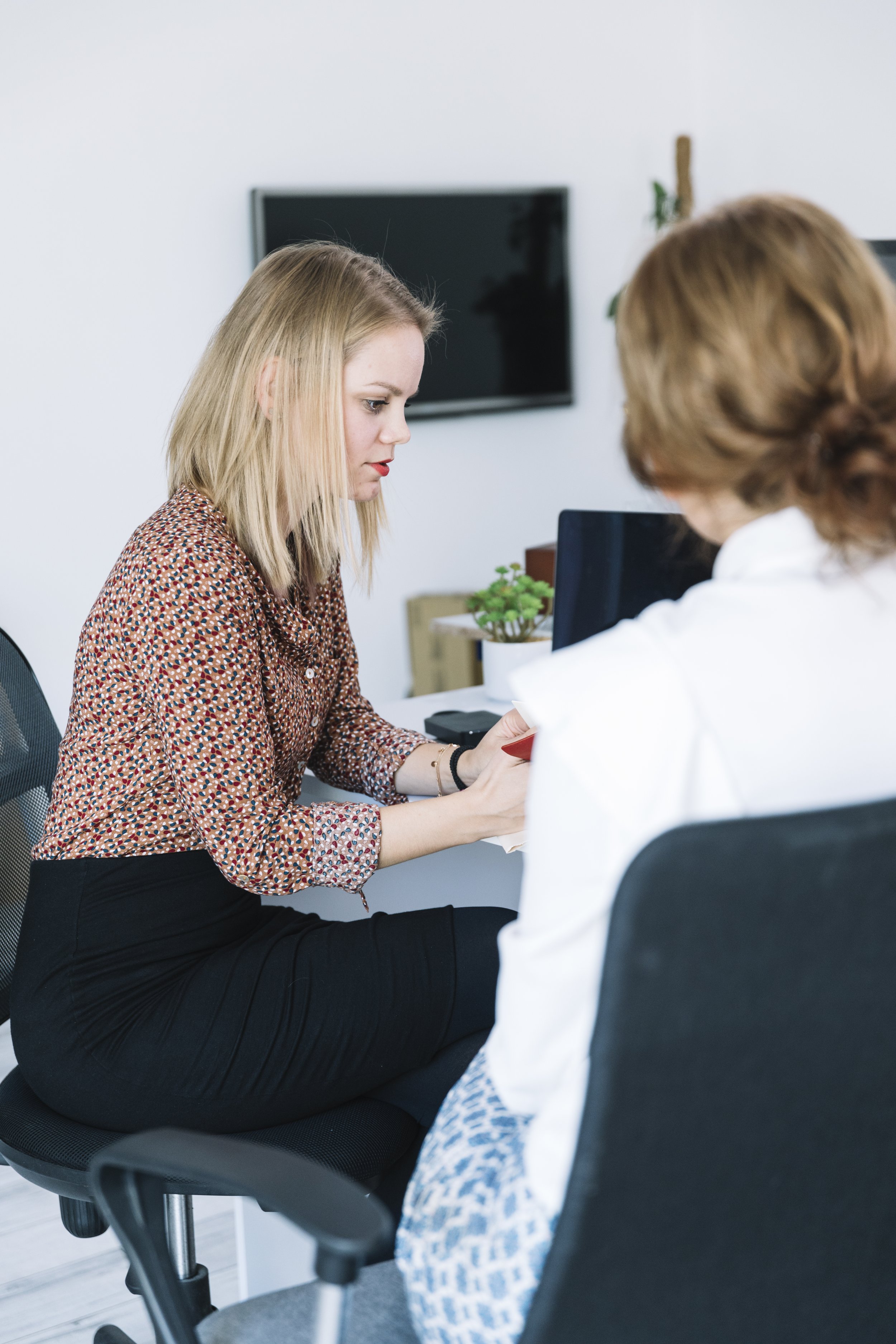 Two women are seated in an office, engaged in a conversation. One woman has blonde hair, wears a patterned blouse, and is listening attentively. The other woman, partially visible, has light brown hair, wears a white top, and is interacting with the first woman. There's a computer monitor and a small green plant on the desk in front of them, with a TV monitor mounted on the wall behind.