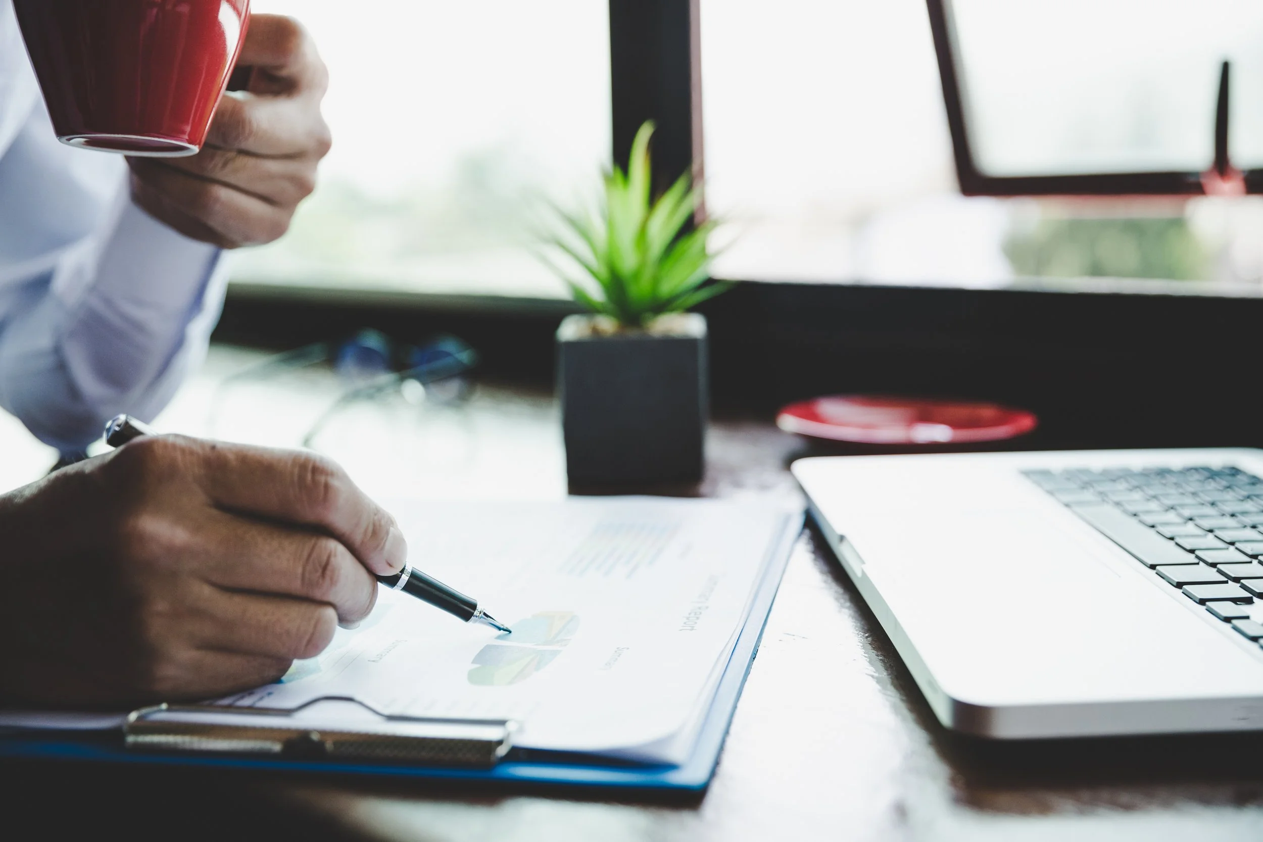 Person holding a red coffee mug and writing on a document on a desk, with a laptop, potted plant, and screen in the background.
