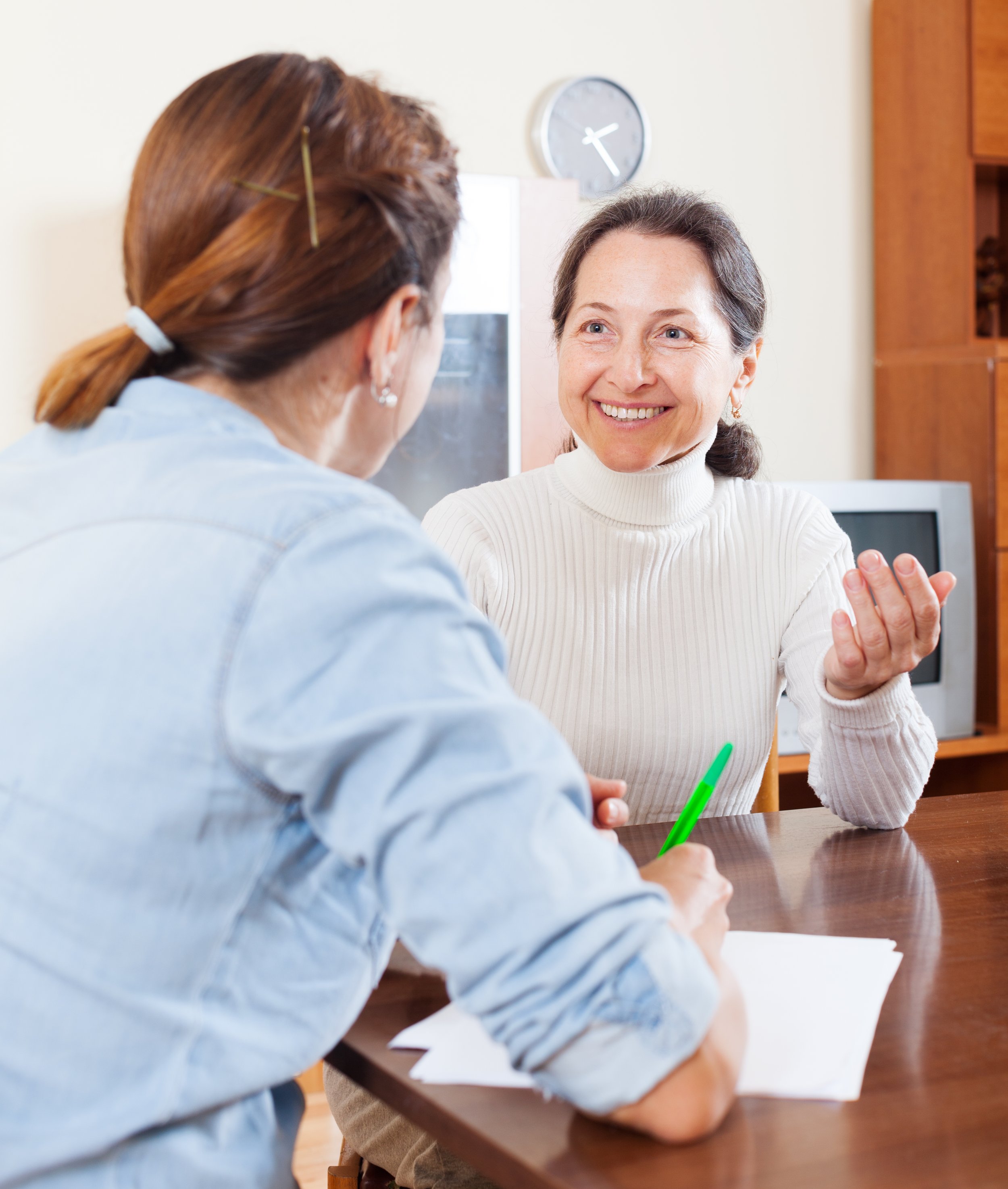 Two women engaging in a friendly conversation across a wooden table, one woman smiling and holding a green pen, the other woman with her back to the camera, in a cozy indoor setting with a wall clock behind them.