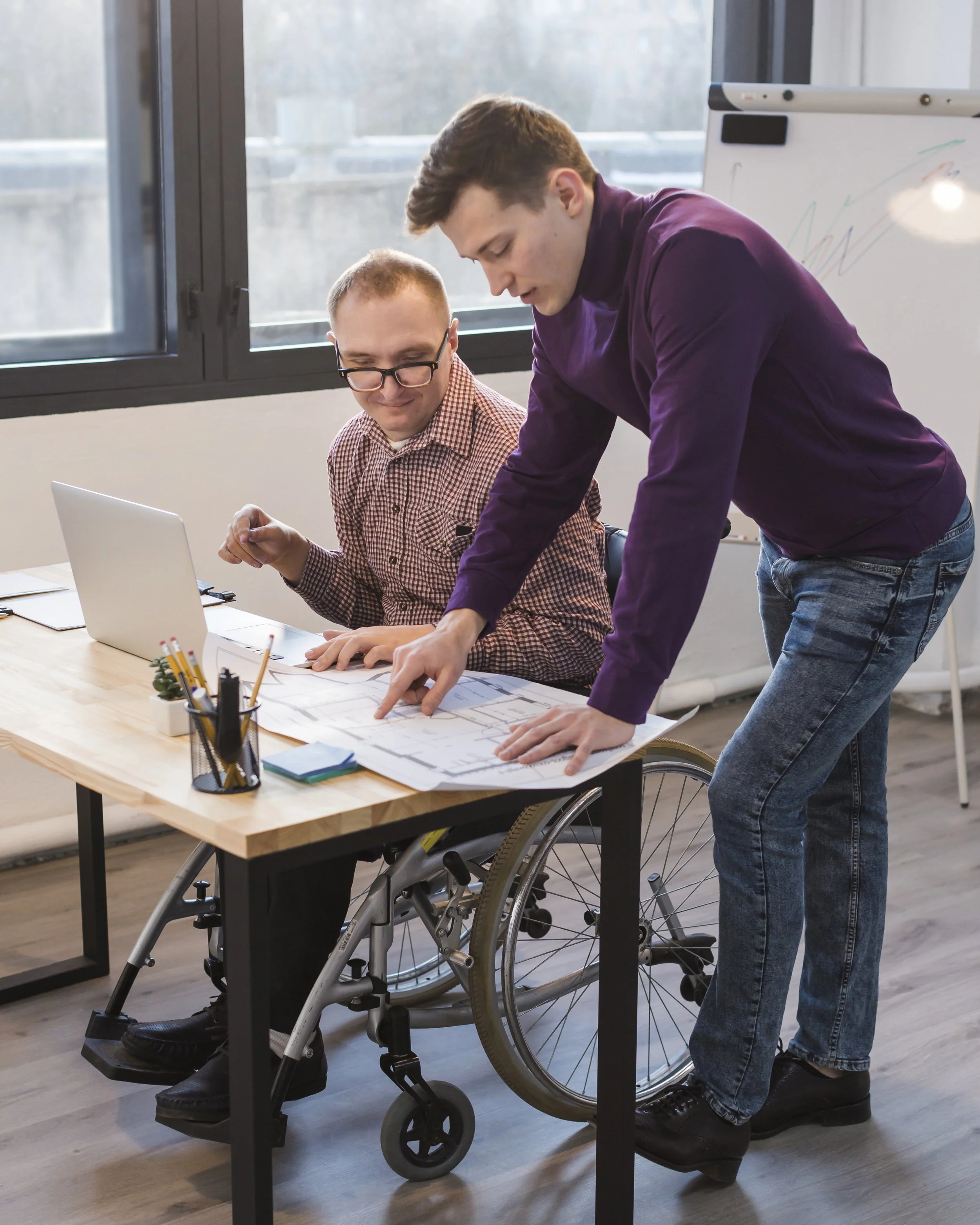 Two men in a modern office, one in a wheelchair, discussing documents on a wooden table. The man in the wheelchair is wearing a checkered shirt and glasses, and the standing man is in a purple shirt and jeans. There is a window in the background and a whiteboard with colorful scribbles.
