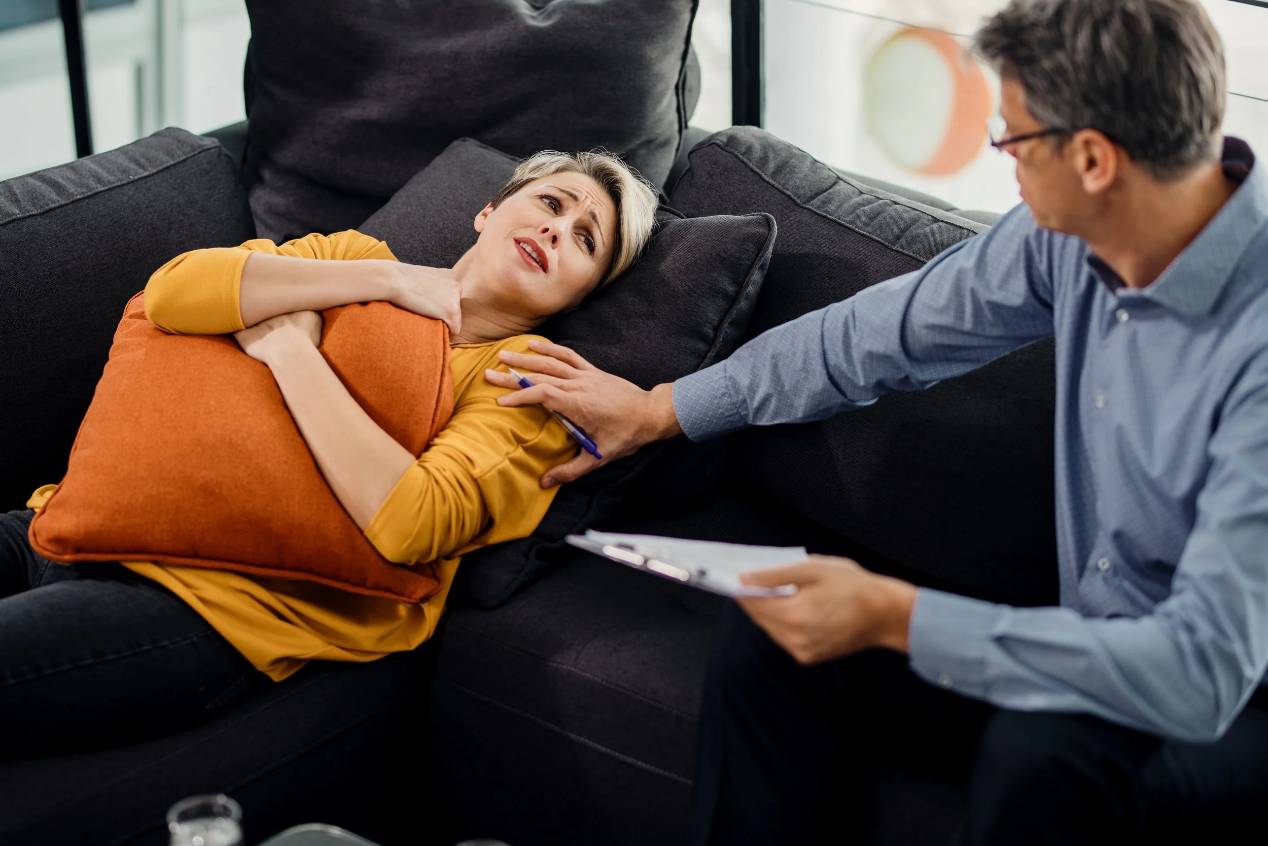 Woman lying on a sofa, appearing distressed, talking with a man who is holding a pen and notebook, while another person stands nearby in a room.