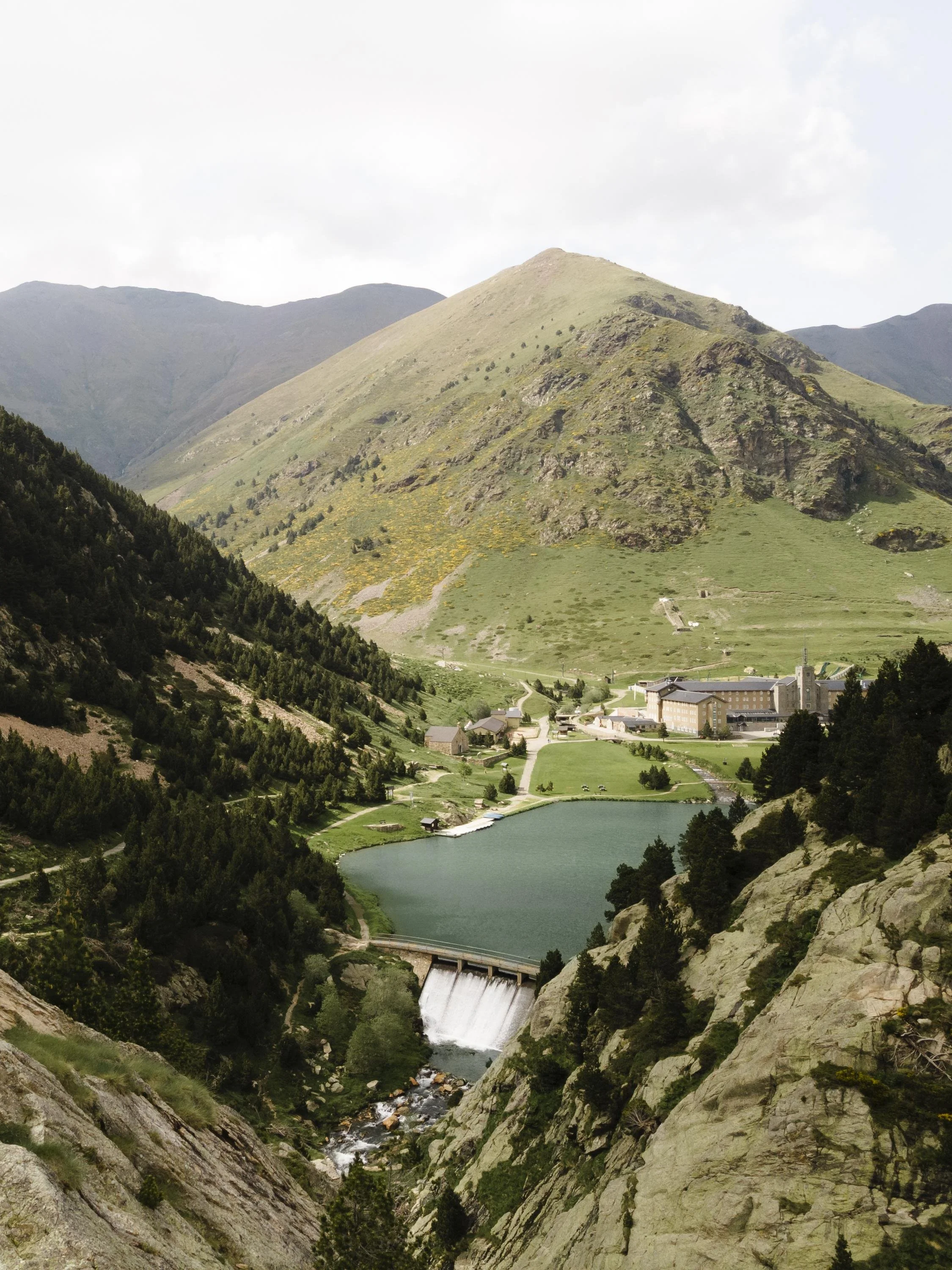 Mountain landscape with a river and waterfall, surrounded by green and rocky hills, a small town and lake at the base, under a cloudy sky.