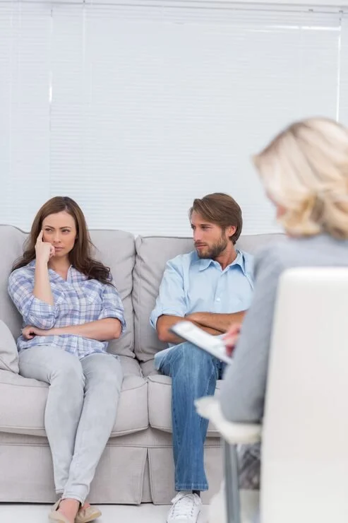 A woman and a man sitting on a couch during a therapy session, talking with a therapist taking notes.