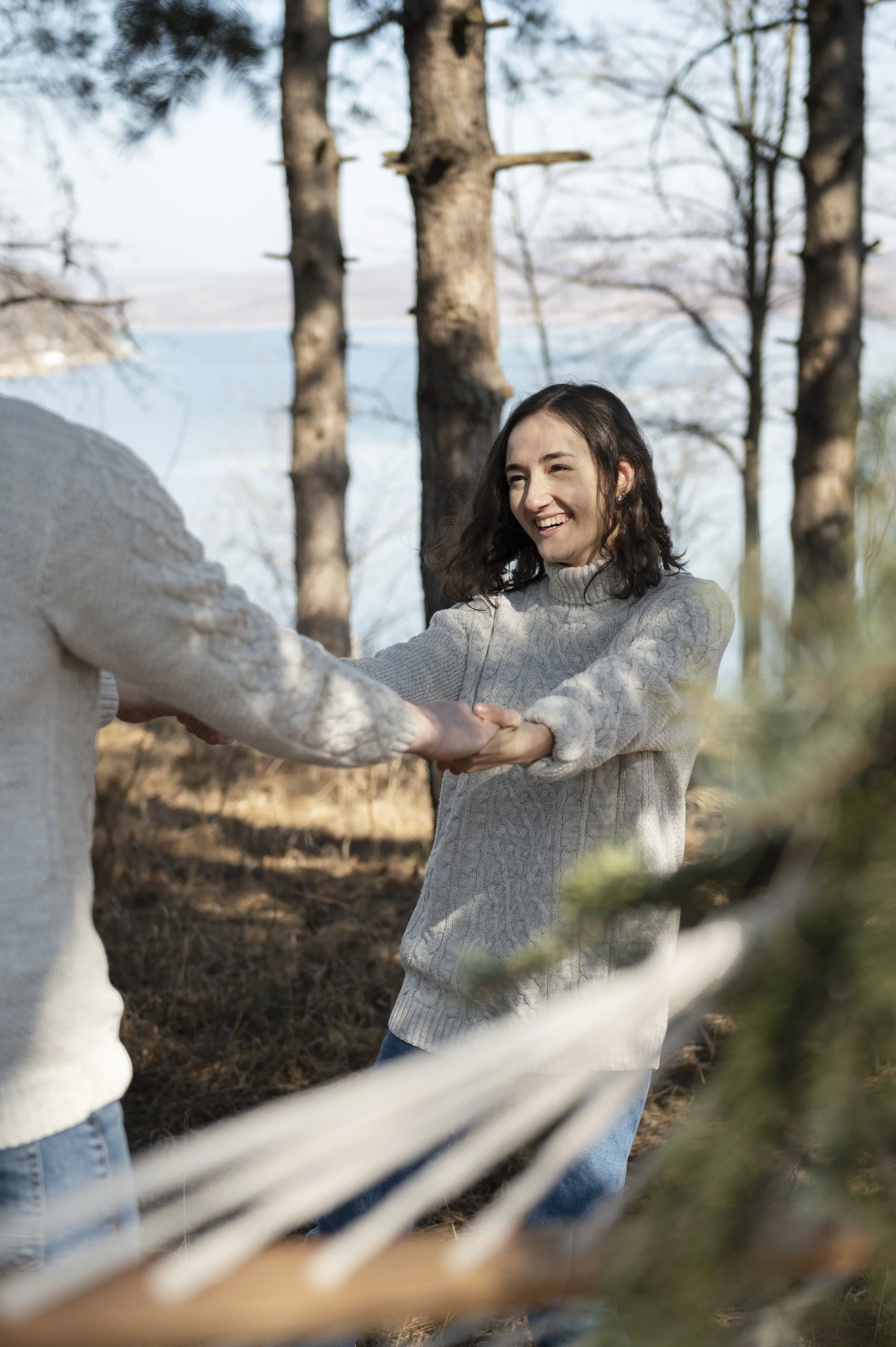 A young woman with curly brown hair, wearing a light gray knitted sweater, smiling and holding hands with someone outside during daytime in a wooded area with tall trees and a lake in the background.