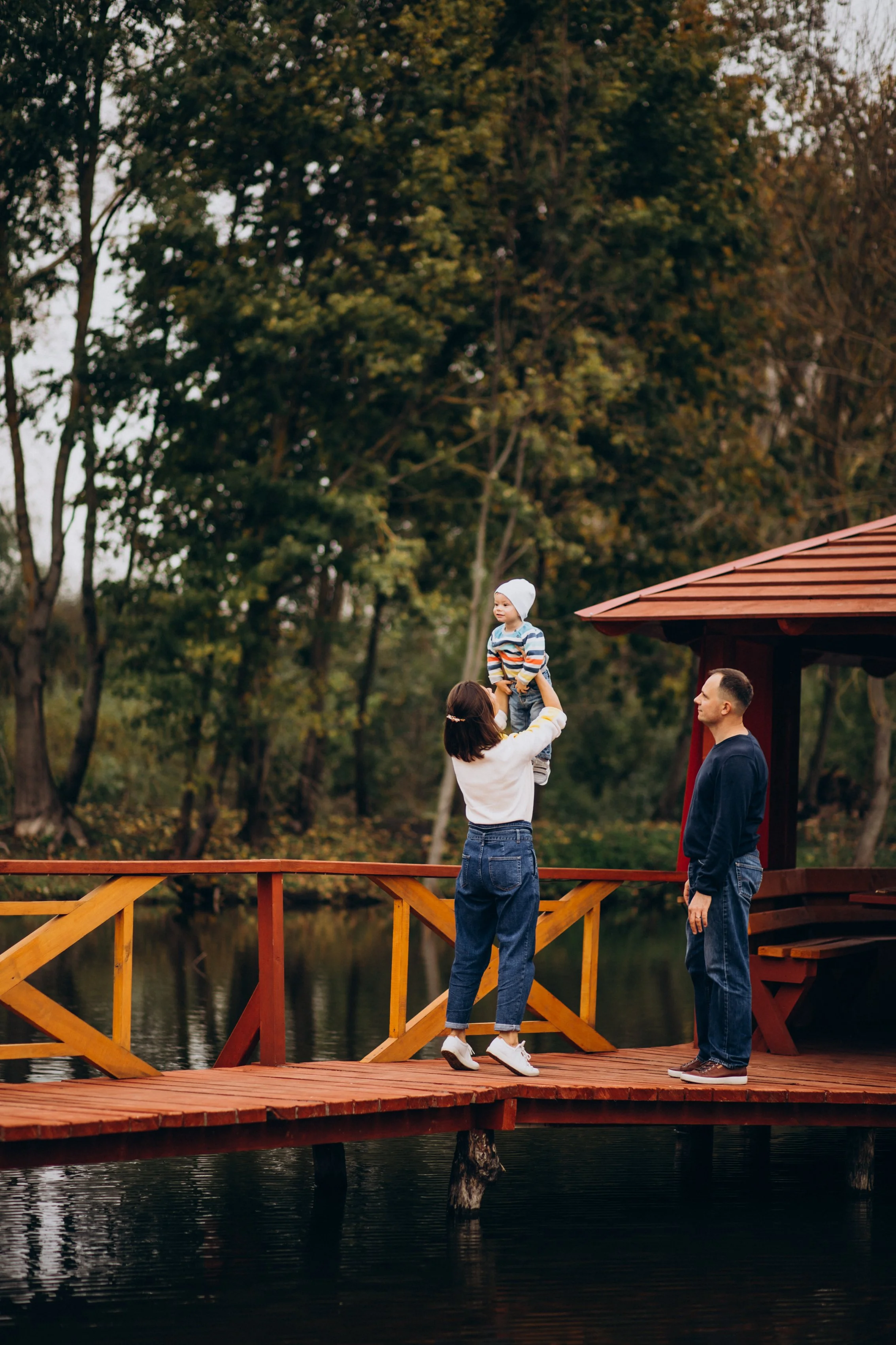 A woman on a wooden deck over water holding a young boy up. The boy is wearing a white beanie, striped sweater, and jeans. A man stands nearby, watching them. The scene is set outdoors with trees in the background.