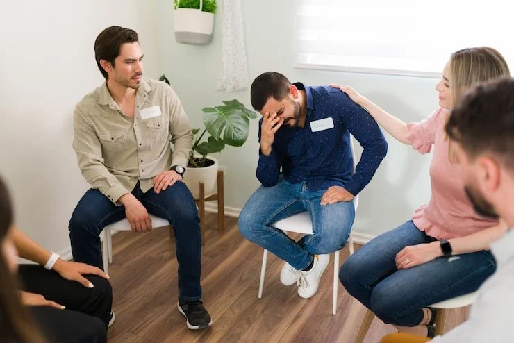 People sitting in a circle in a therapy or support group setting, offering comfort to a man with his hand on his face, expressing distress.