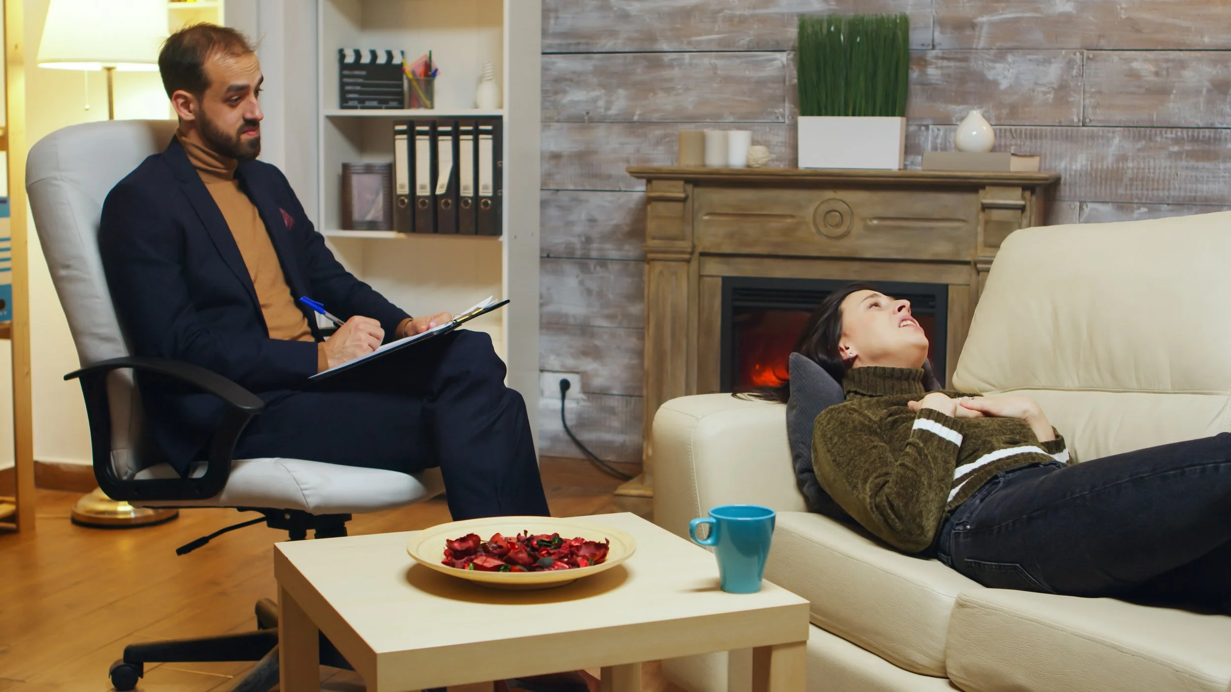 A man in a suit sitting on a white chair, holding a clipboard and pen, looking at a woman lying on a sofa with her head tilted back, smiling. The woman is wearing a brown sweater with white stripes on the sleeves, and the room has a cozy, modern decor with a wooden fireplace, candles, and a bookshelf.