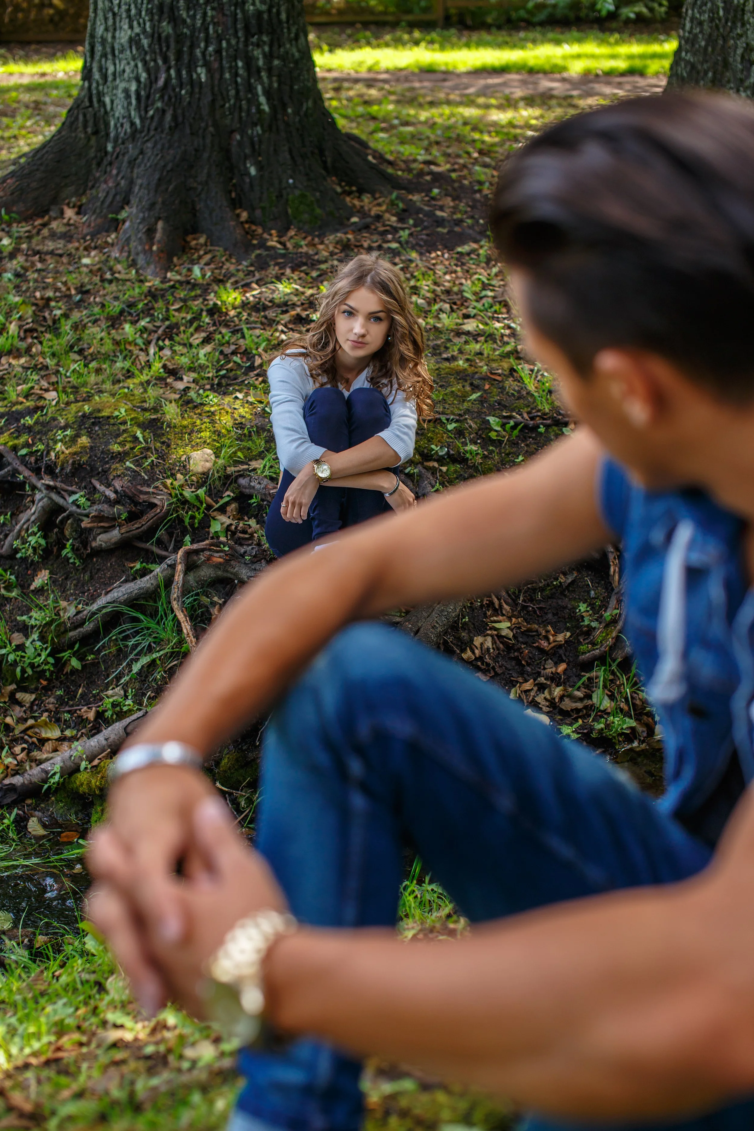 A woman with curly hair sitting on the grass near a large tree, looking directly at the camera with a serious expression. A person in the foreground, slightly out of focus, appears to be holding hands or touching the woman's arm. The scene is outdoors in a wooded area with green grass and sunlight filtering through the trees.