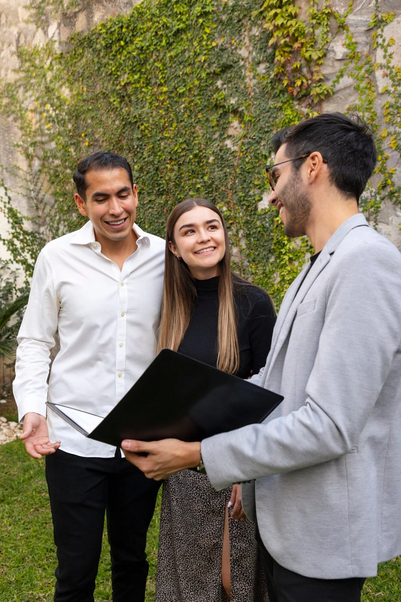 Three people standing outdoors in front of a vine-covered stone wall, engaged in conversation and smiling, with one holding a black folder or notebook.