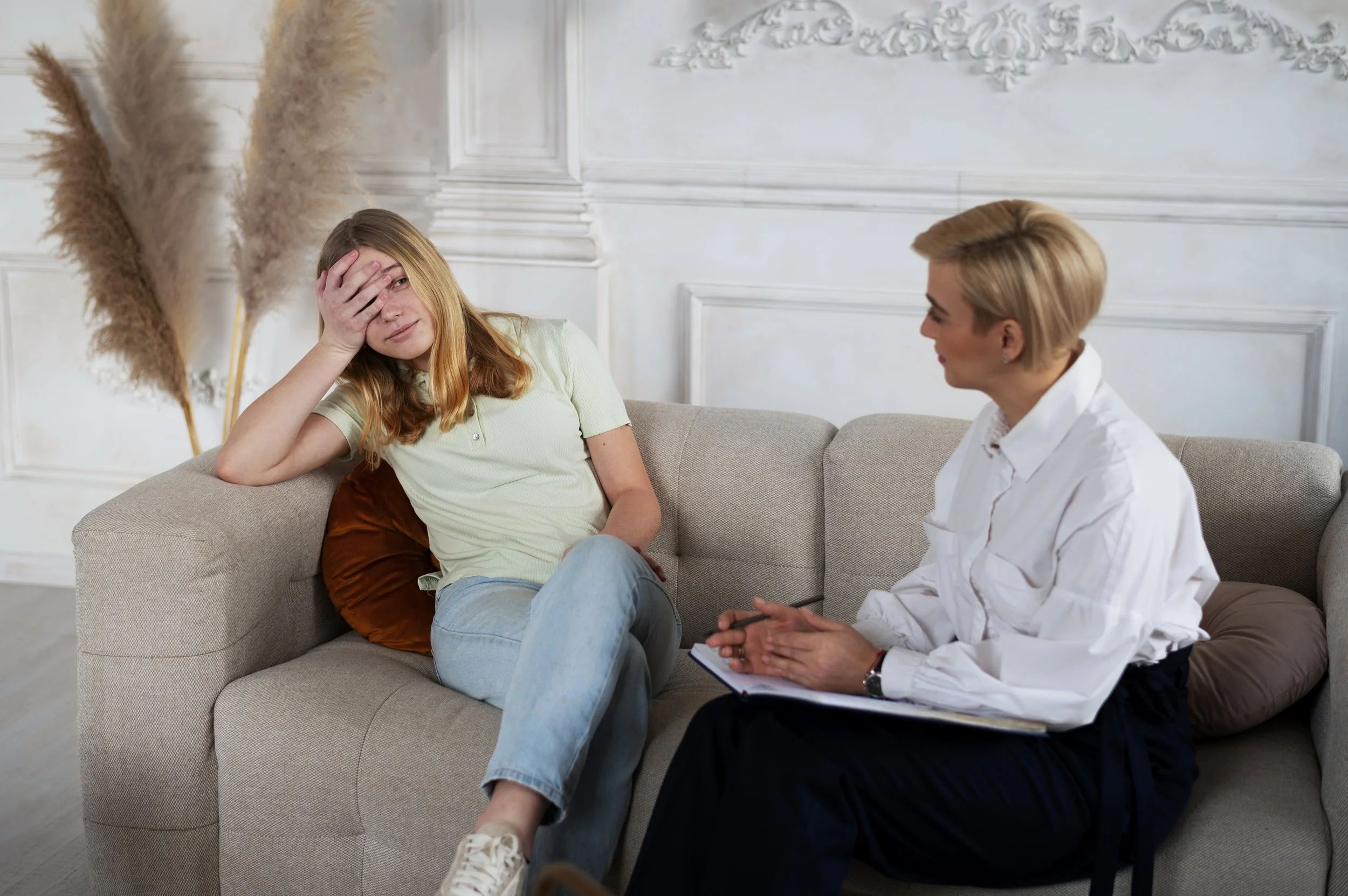 A woman sitting on a beige couch with her hand on her forehead, appearing distressed, while another woman, possibly a therapist or counselor, listens and takes notes in a notebook during a conversation in a bright, tastefully decorated room.