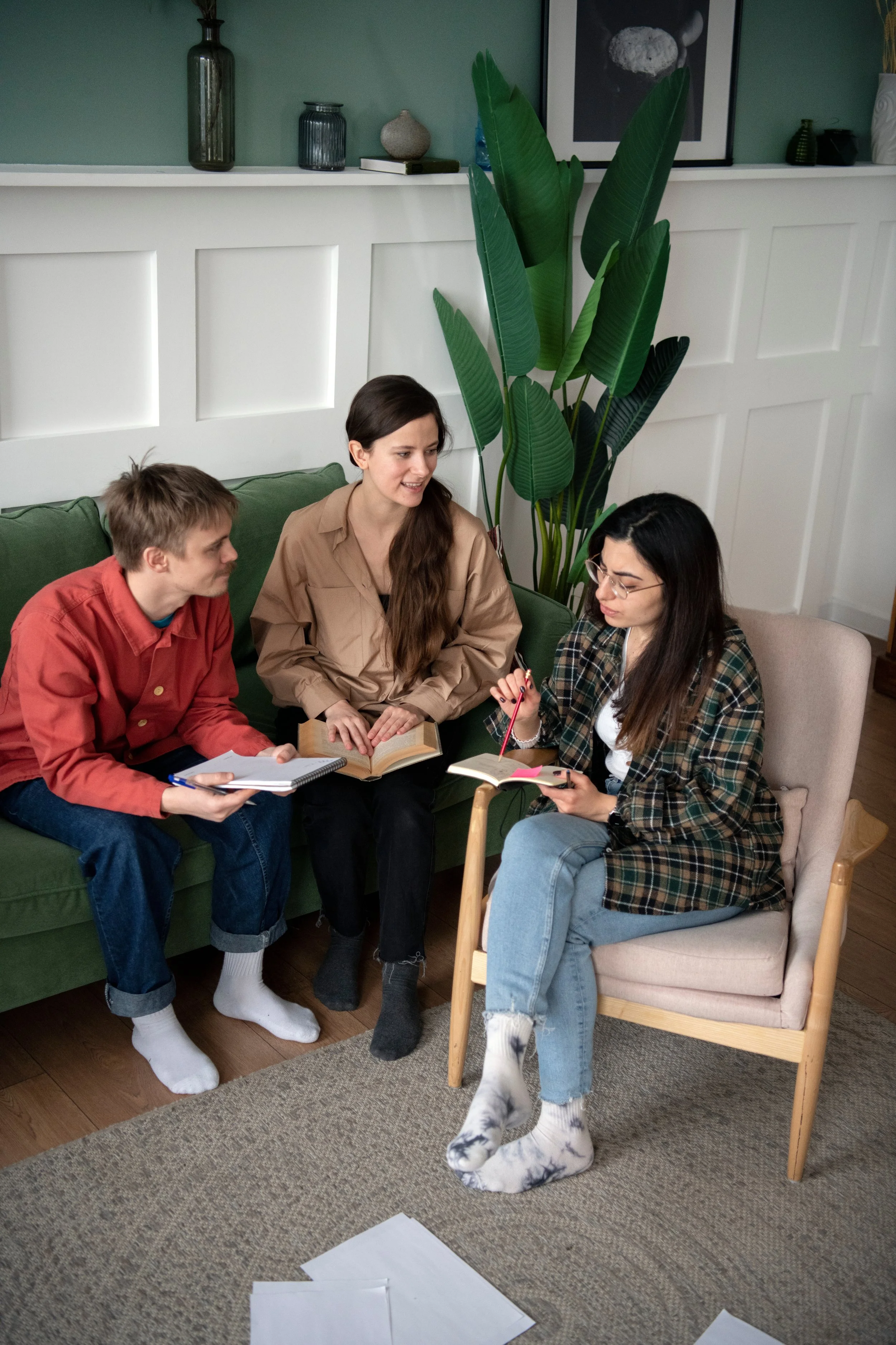 Three young women sitting together in a living room, engaged in conversation. One is holding a notebook with papers, the second has an open book, and the third is holding a notepad and pen. They are seated on a green sofa and a beige armchair, wearing casual clothes and socks, with scattered papers on the carpet in front of them. A large houseplant and framed artwork are in the background.