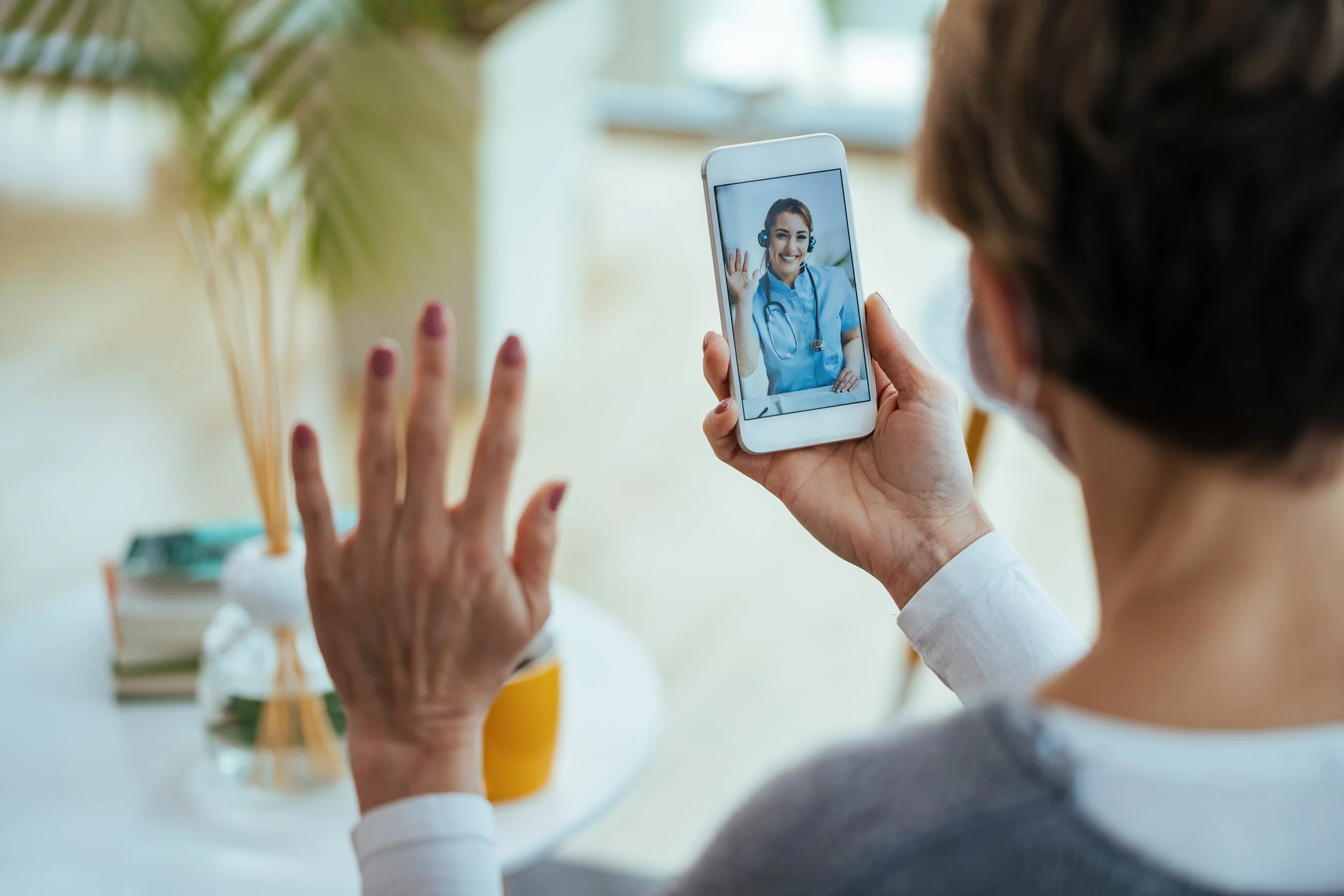 A person holding a smartphone takes a selfie of a female healthcare worker wearing scrubs and a stethoscope, smiling and waving in a bright room with houseplants in the background.