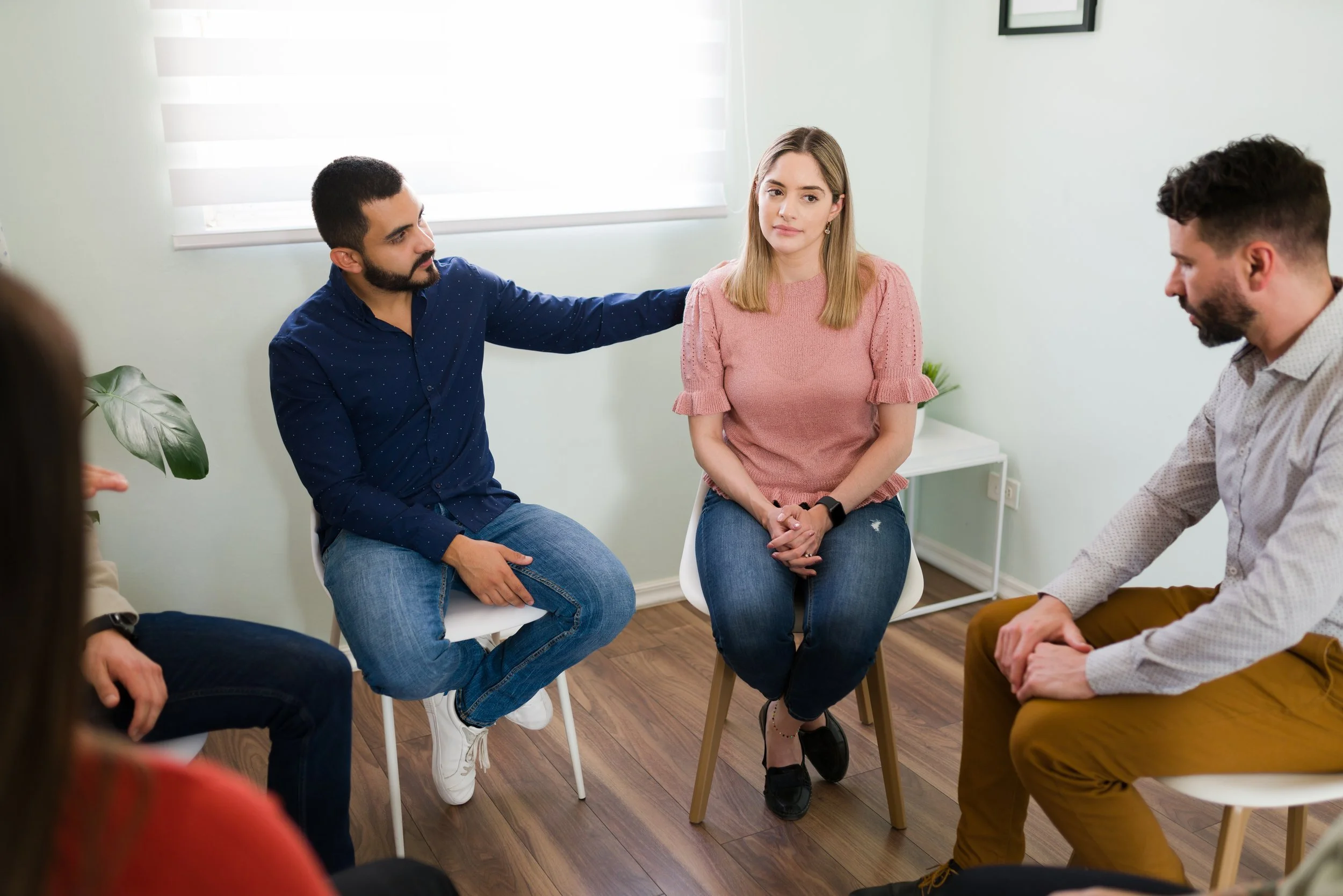 A group therapy session with five people sitting in a circle, including a woman in a pink sweater and men in casual clothing, in a bright room with light-colored walls and a plant.