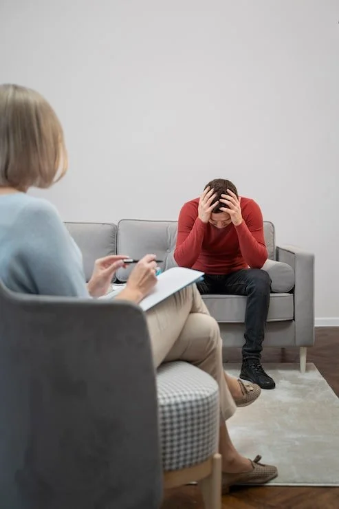 A man sits on a couch with his head in his hands, appearing distressed, while a therapist takes notes during a therapy session in a minimalist office.