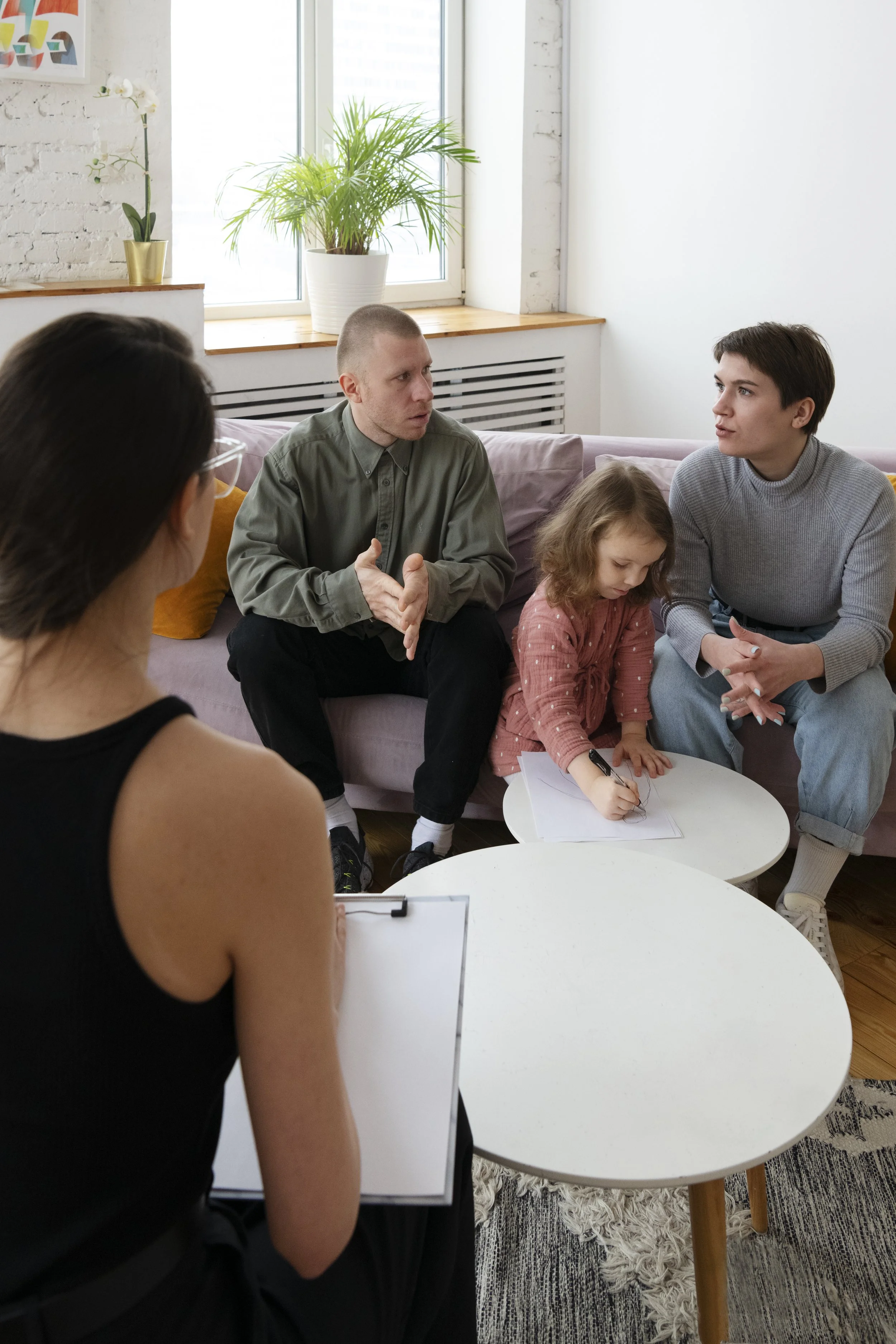 A group of people in a living room having a discussion, including a woman with glasses in the foreground, two men on a sofa, and a young girl writing or drawing at a small white table.