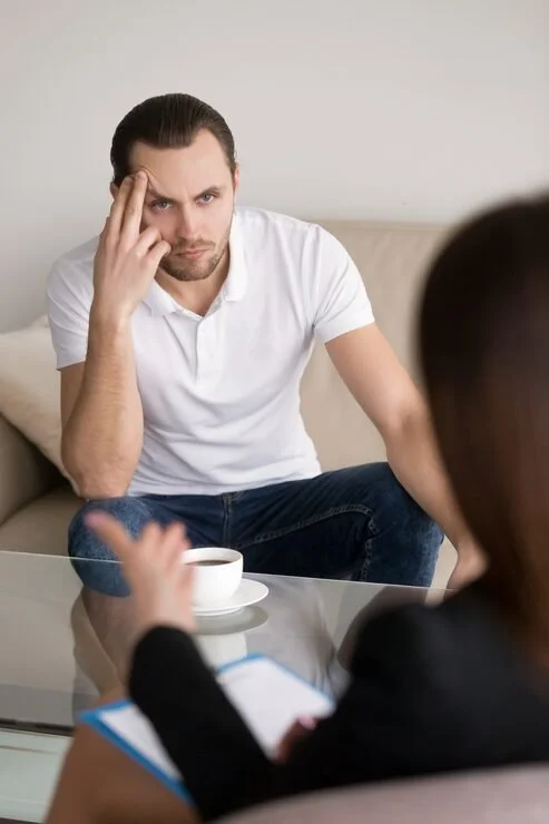 Man dressed in white shirt sitting on a sofa, appearing concerned, during a therapy or counseling session with a woman taking notes.