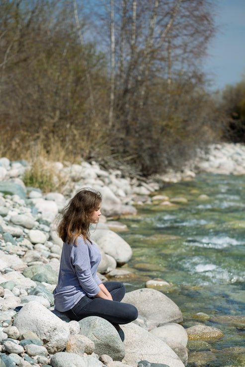 A woman with long brown hair sitting on large rocks by a river, wearing a light purple shirt and black pants, with trees in the background and a partly cloudy sky.