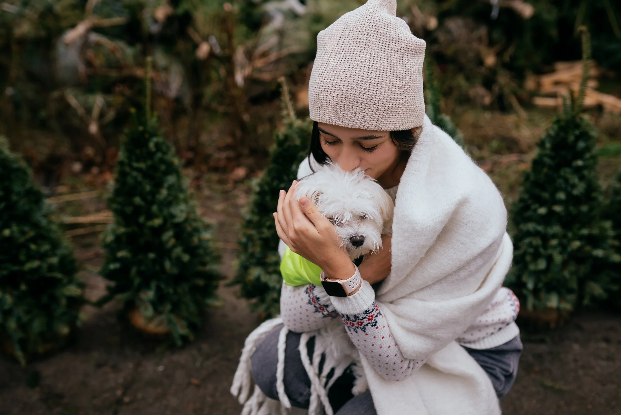 A woman wearing a beige knit hat and white sweater hugging a small white fluffy dog outdoors in a garden with green plants.