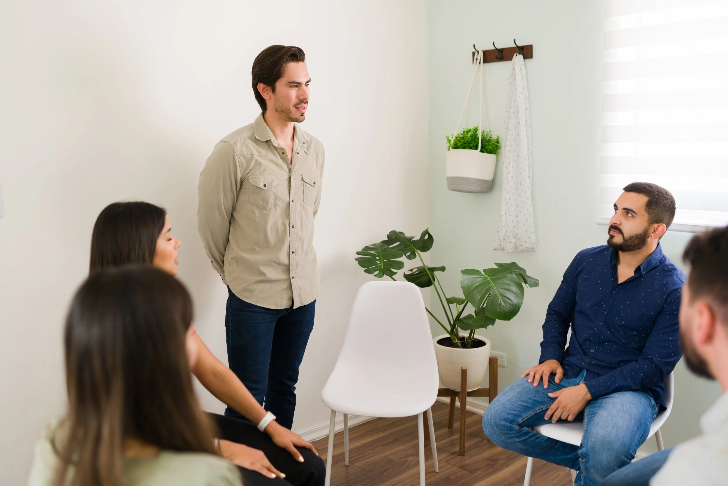 A man with dark hair and a beard standing and speaking to a group of seated people in a room with white walls, large windows, and houseplants.