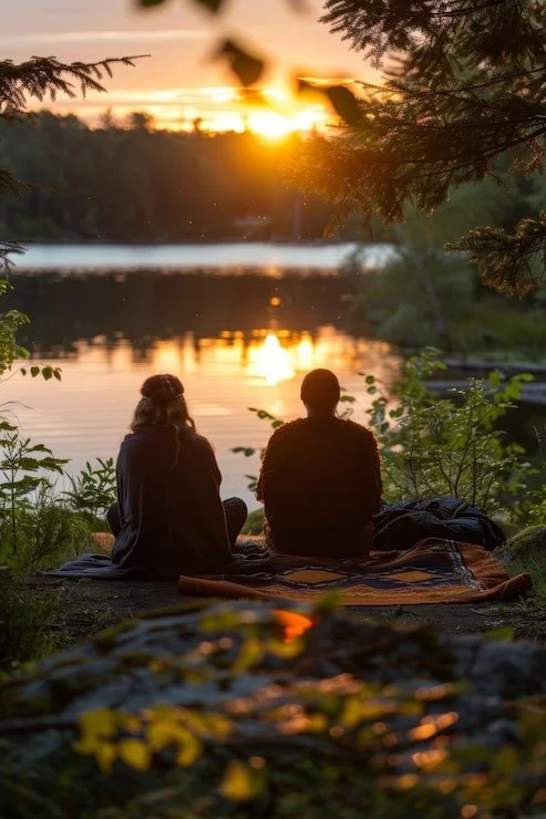 Two people sitting on a blanket by a peaceful lake during sunset, surrounded by trees and nature.