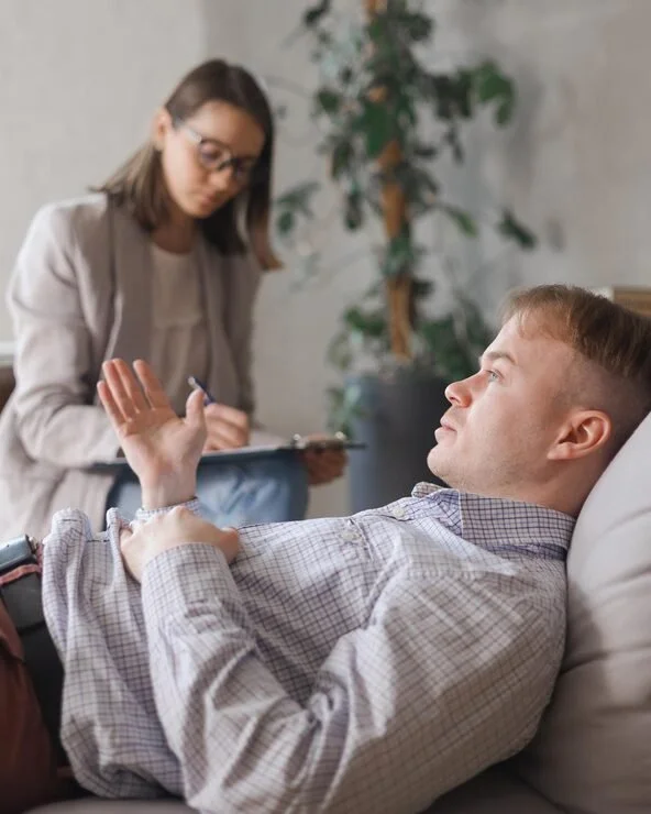 A man lying on a sofa, gesturing with his right hand, while a woman standing nearby appears to be taking notes or filling out a form, in a room with a plant and a gray wall.